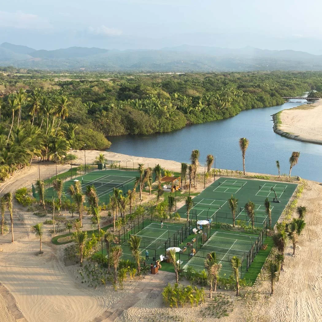 Aerial view of a tropical resort with multiple tennis courts surrounded by palm trees, a beach, a river, and lush green landscape with mountains in the background.