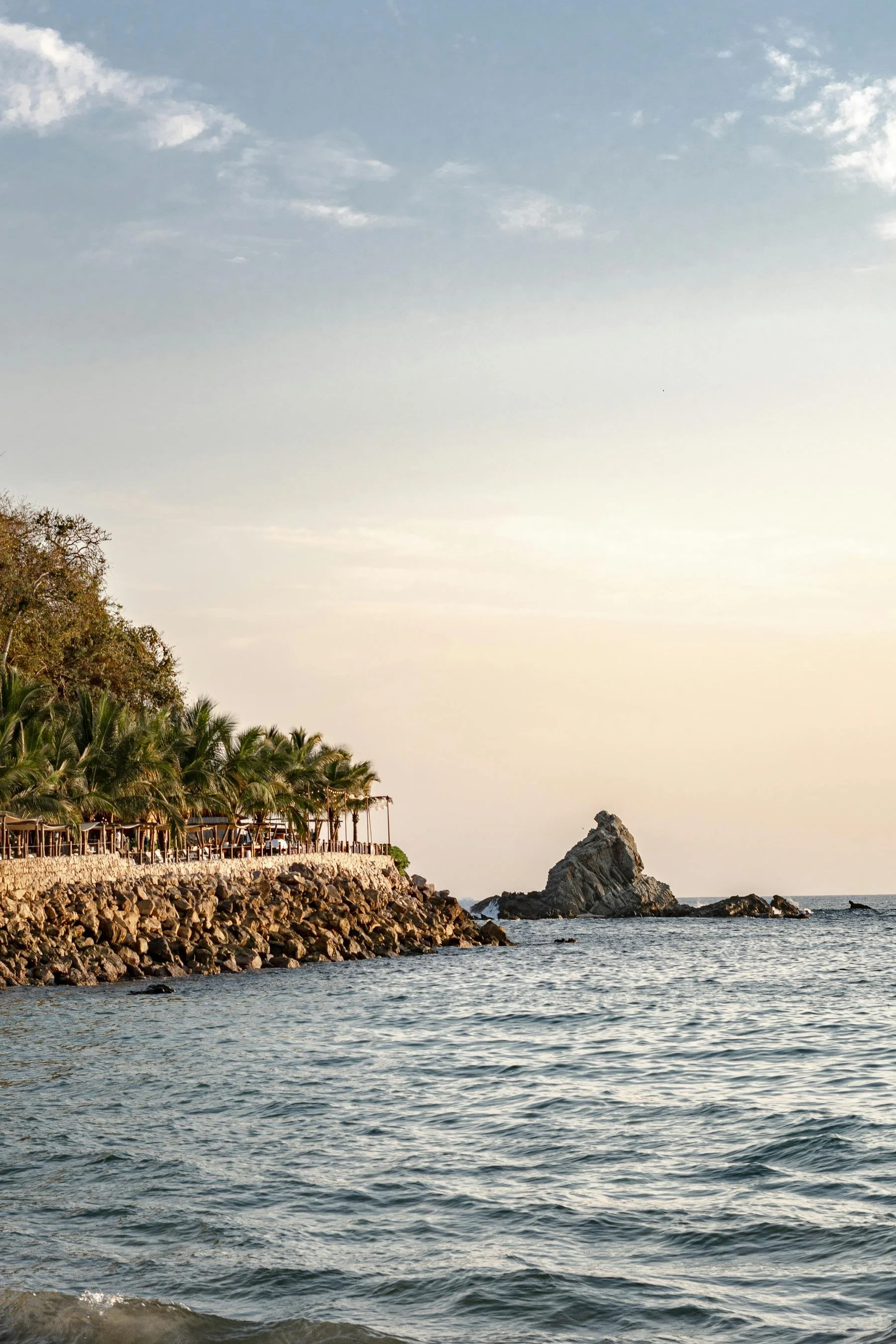 A coastal scene with a rocky pier, palm trees, and a rock formation in the water, under a partly cloudy sky during sunset.
