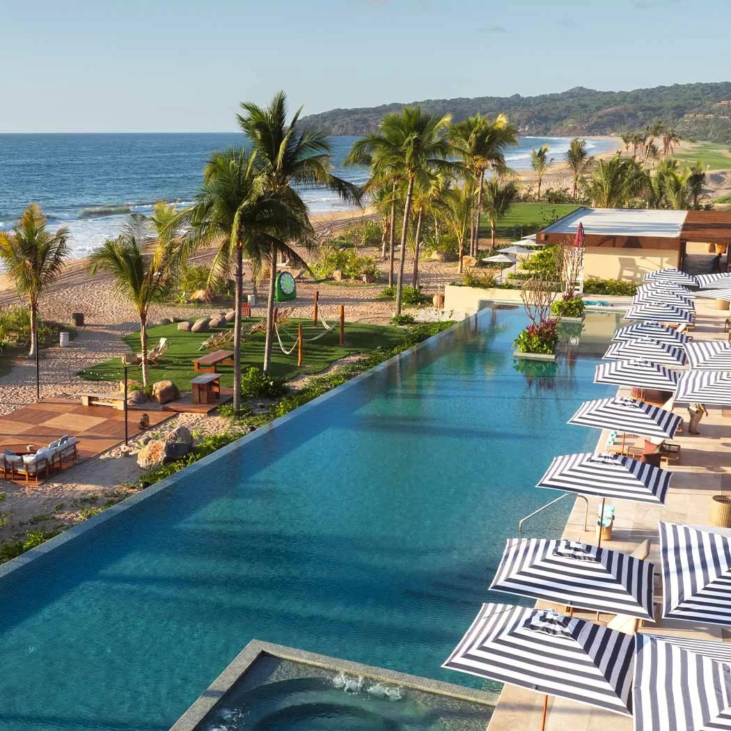 A luxurious resort with an infinity pool overlooking a beach and ocean, lined with striped umbrellas and palm trees.