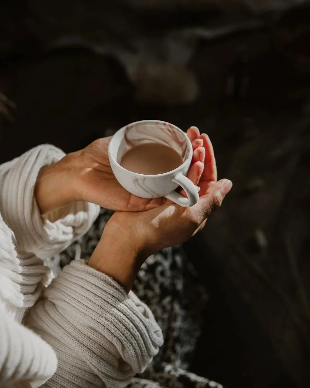 Person wearing a beige sweater holding a white mug with a marble pattern, filled with coffee or tea, over a dark background.