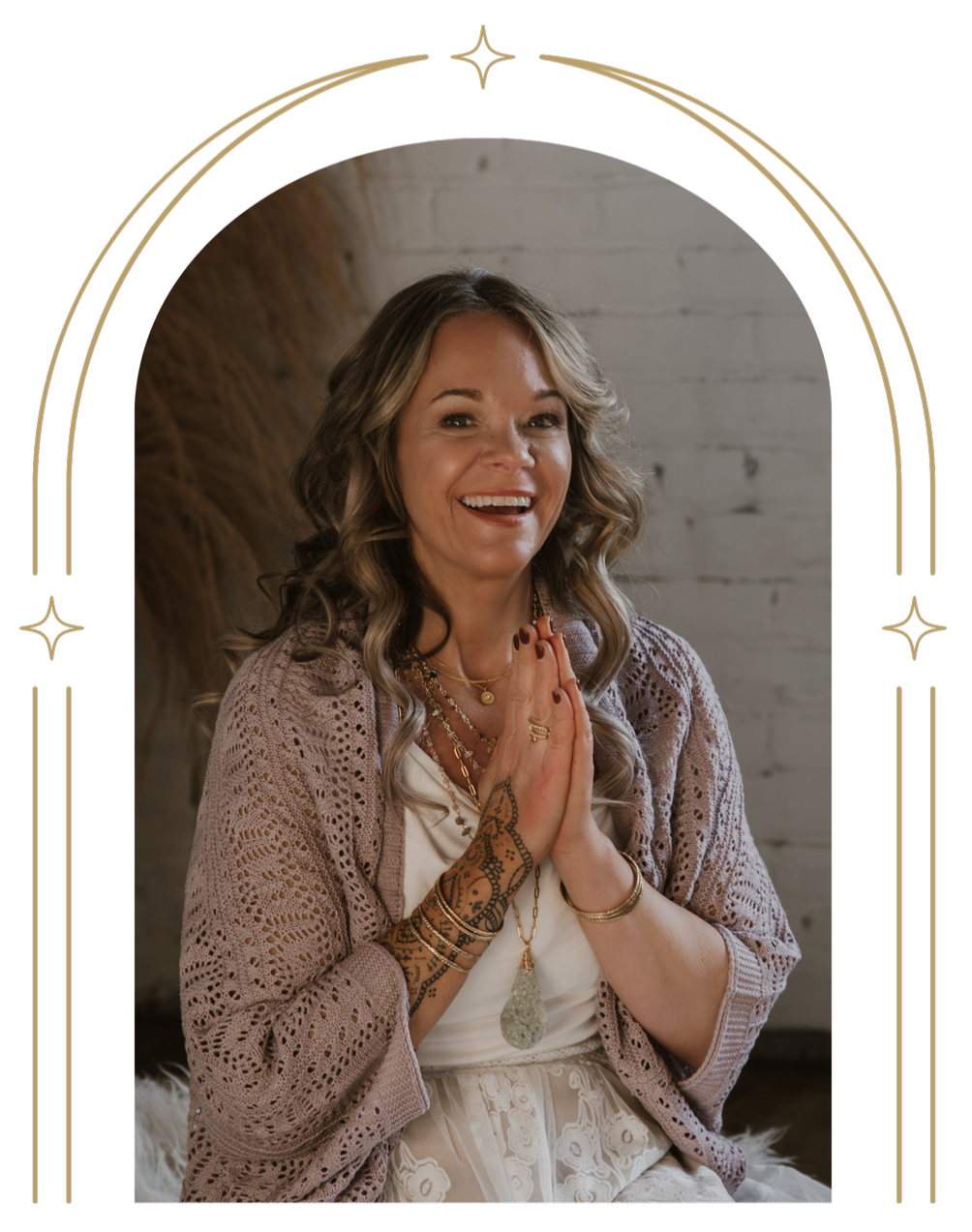 A smiling woman with wavy blonde hair and tattoos on her right arm, wearing a light beige crochet cardigan and multiple gold necklaces, sitting with her hands in a prayer pose against a rustic brick wall background.