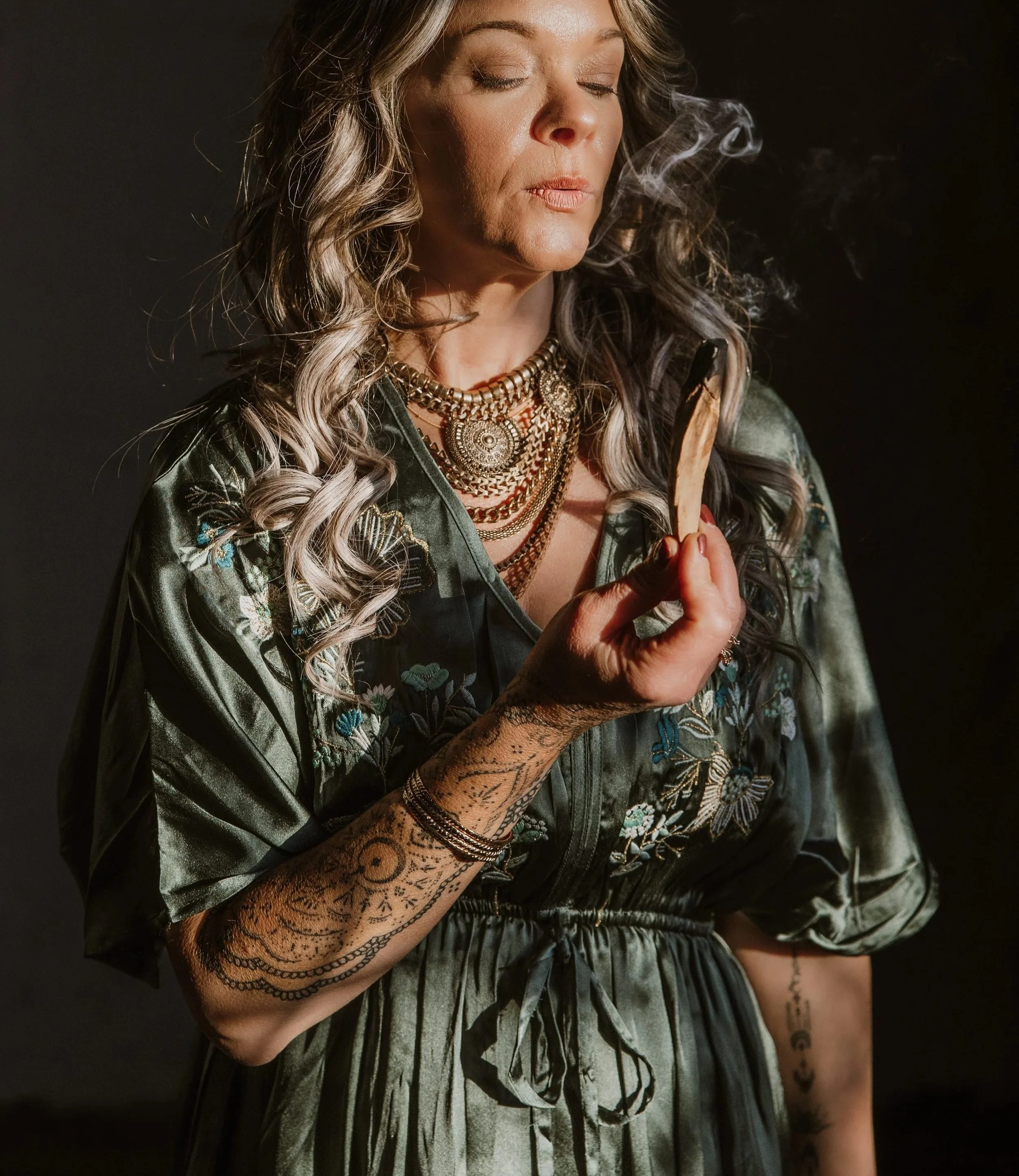 A woman with wavy blonde hair and tattoos on her arm holding a burning cigarette or joint, wearing layered necklaces and a satin embroidered jacket, with smoke swirling near her face against a dark background.