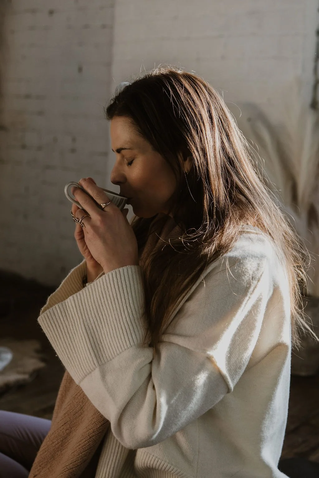 A woman with long brown hair wearing a cream-colored sweater, holding a mug close to her face with her eyes closed, enjoying a warm beverage in a cozy, softly lit room.