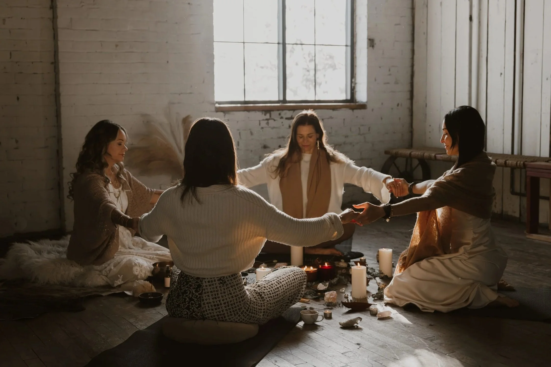 Group of women sitting in a circle holding hands, participating in a spiritual or meditation session in a rustic, well-lit room with candles and candles on the floor.