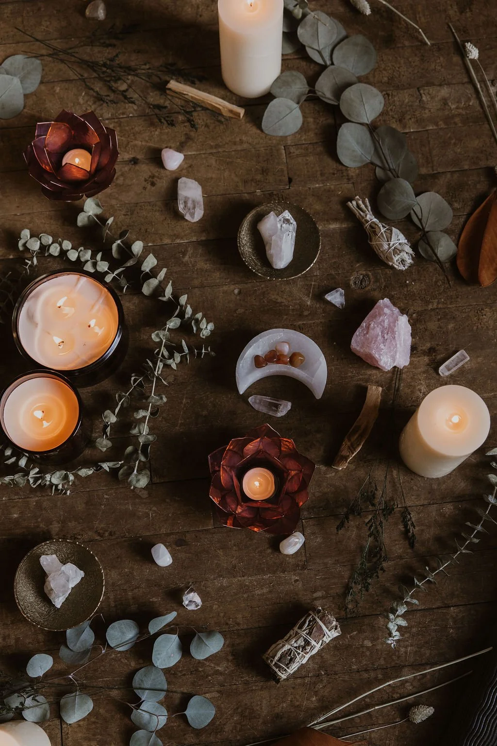 A rustic wooden table decorated with lit candles, eucalyptus leaves, crystals, and small decorative objects, creating a calming and cozy ambiance.