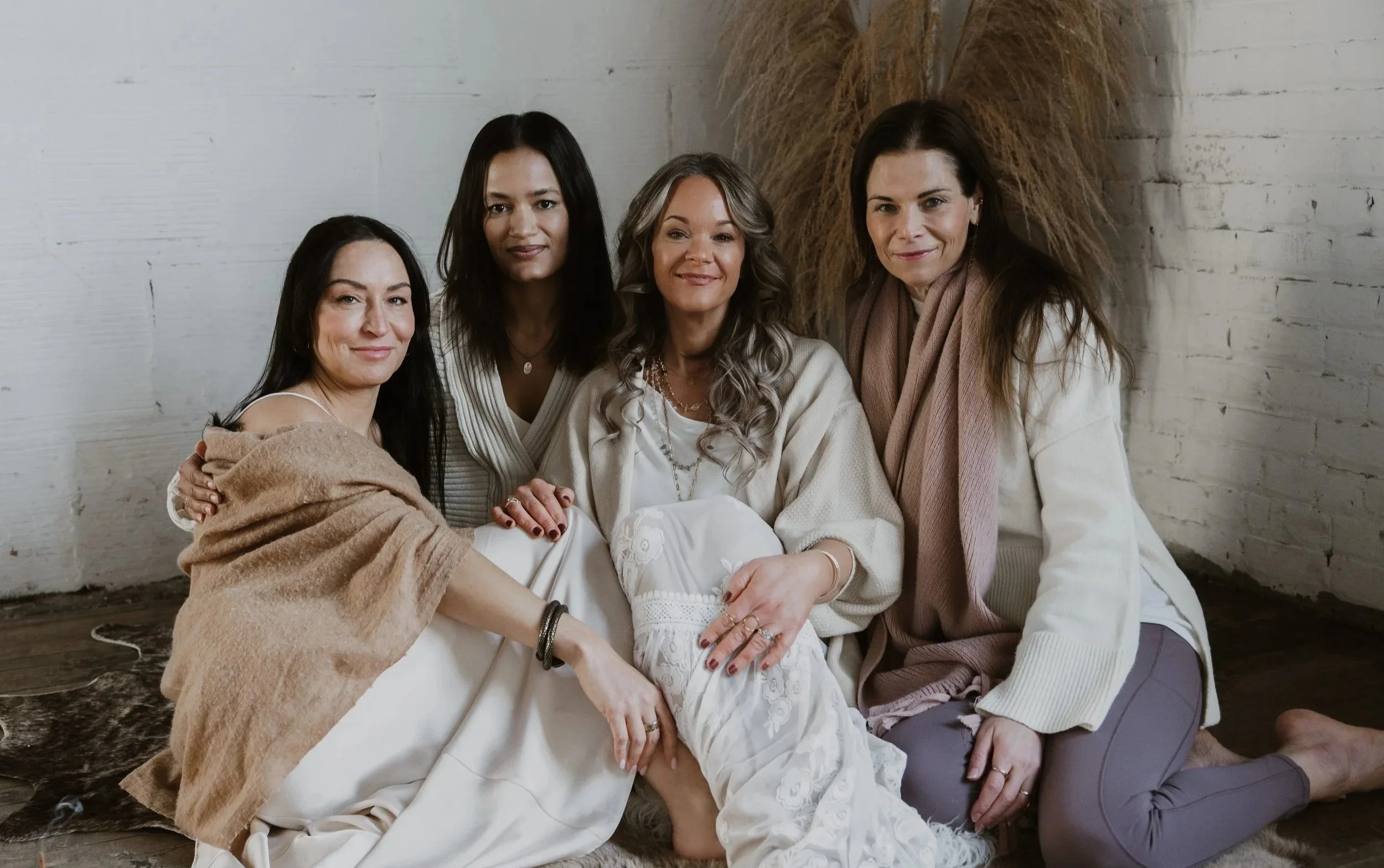 Five women sitting together inside a room with white brick walls, dressed in cozy neutral-toned clothing, smiling warmly at the camera.