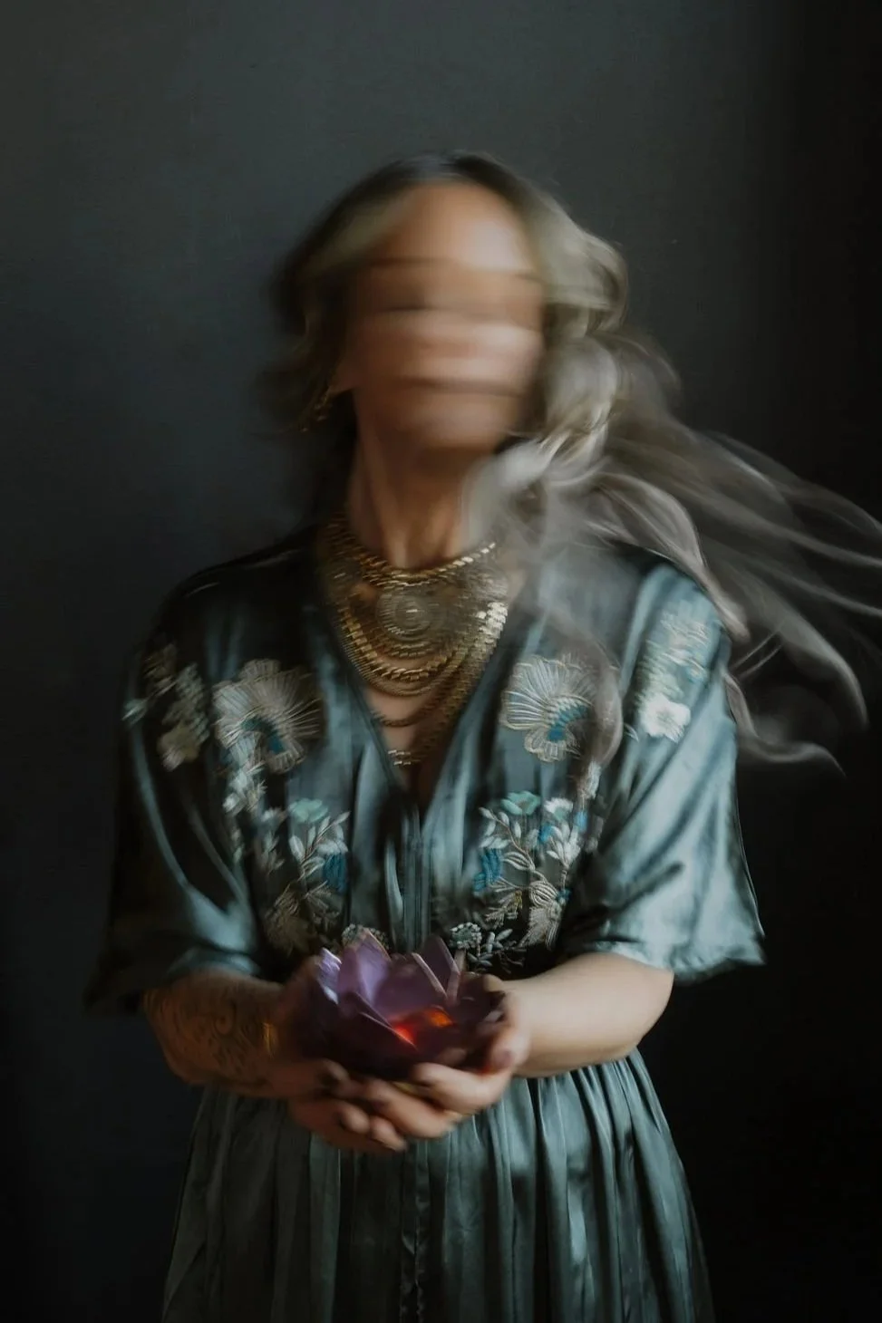 A woman with long, flowing hair in motion, wearing a blue floral dress with embroidery and layered necklaces, holding a purple gemstone.