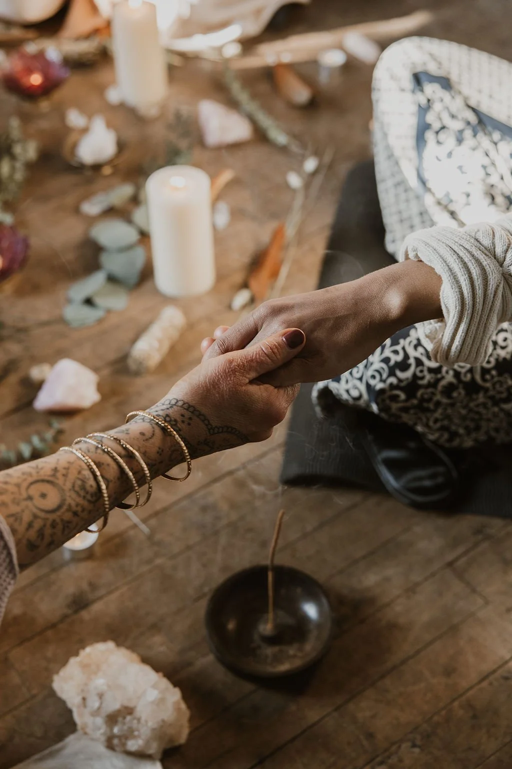 Two people holding hands over a table with rocks, candles, and crystals, indicating a spiritual or ritual setting.