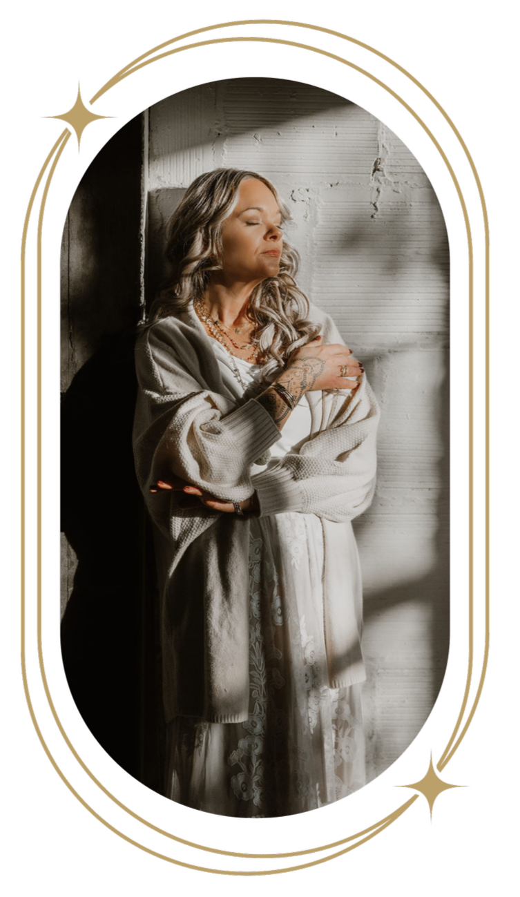 A woman with long, wavy gray hair and tattoos on her hands is standing with her arms crossed and eyes closed, leaning against a textured wall with a shadow cast on her face. She is wearing a beige cardigan and a lace dress, with layered necklaces.