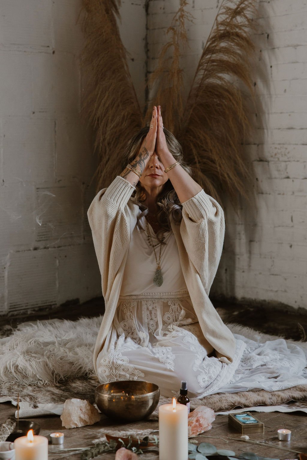A woman in bohemian attire practicing meditation or prayer indoors, sitting cross-legged on a sheepskin rug, with candles, crystals, and incense around her, and decorative pampas grass in the background.