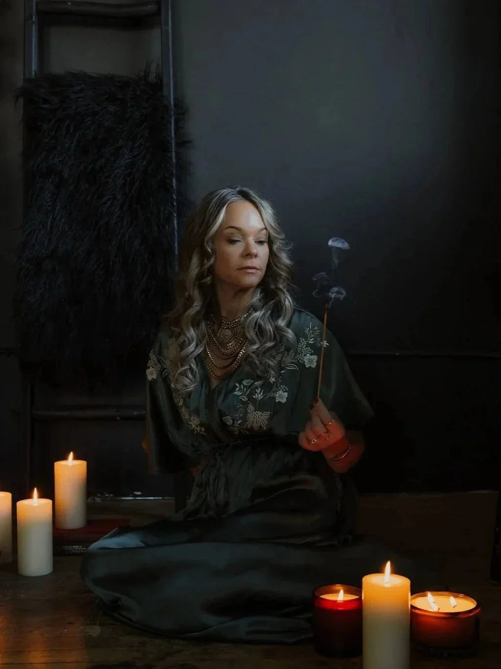 A woman sitting on the floor in a dark room surrounded by lit candles, holding a lit incense stick, with a calm and reflective expression.