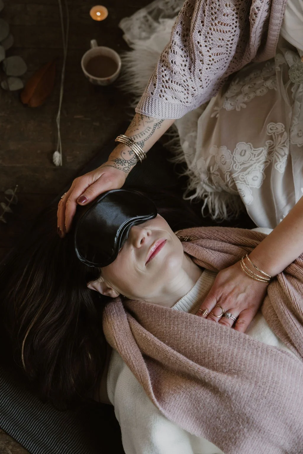 A woman lying down with a blindfold over her eyes, while another woman gently places her hand on her head. The woman lying down appears relaxed with a slight smile, and the scene is set indoors with warm lighting, a cup of tea, and some cozy items nearby.