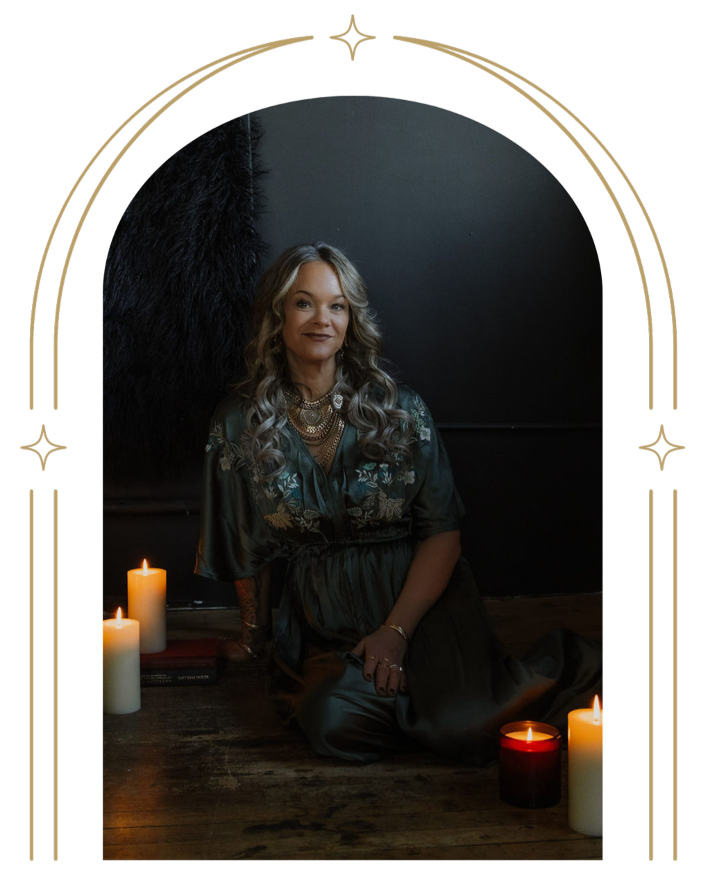 A woman with long curly hair, sitting on a wooden floor surrounded by lit candles, wearing a dark embroidered dress and statement jewelry, in a dimly lit room with a black background.