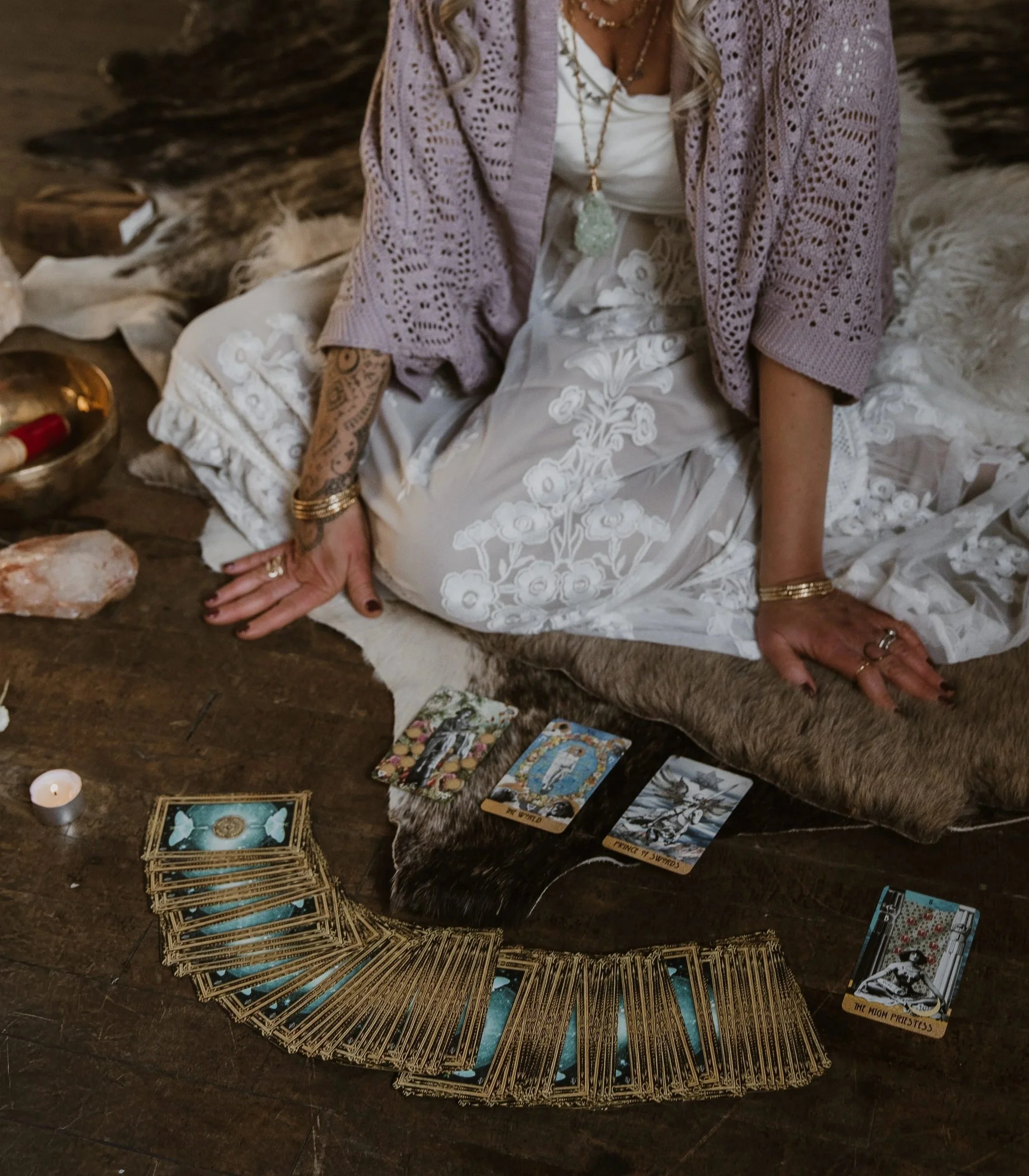 The image shows a woman sitting on a wooden floor surrounded by tarot cards, crystals, a candle, and other spiritual items, with a mystical and spiritual ambiance.