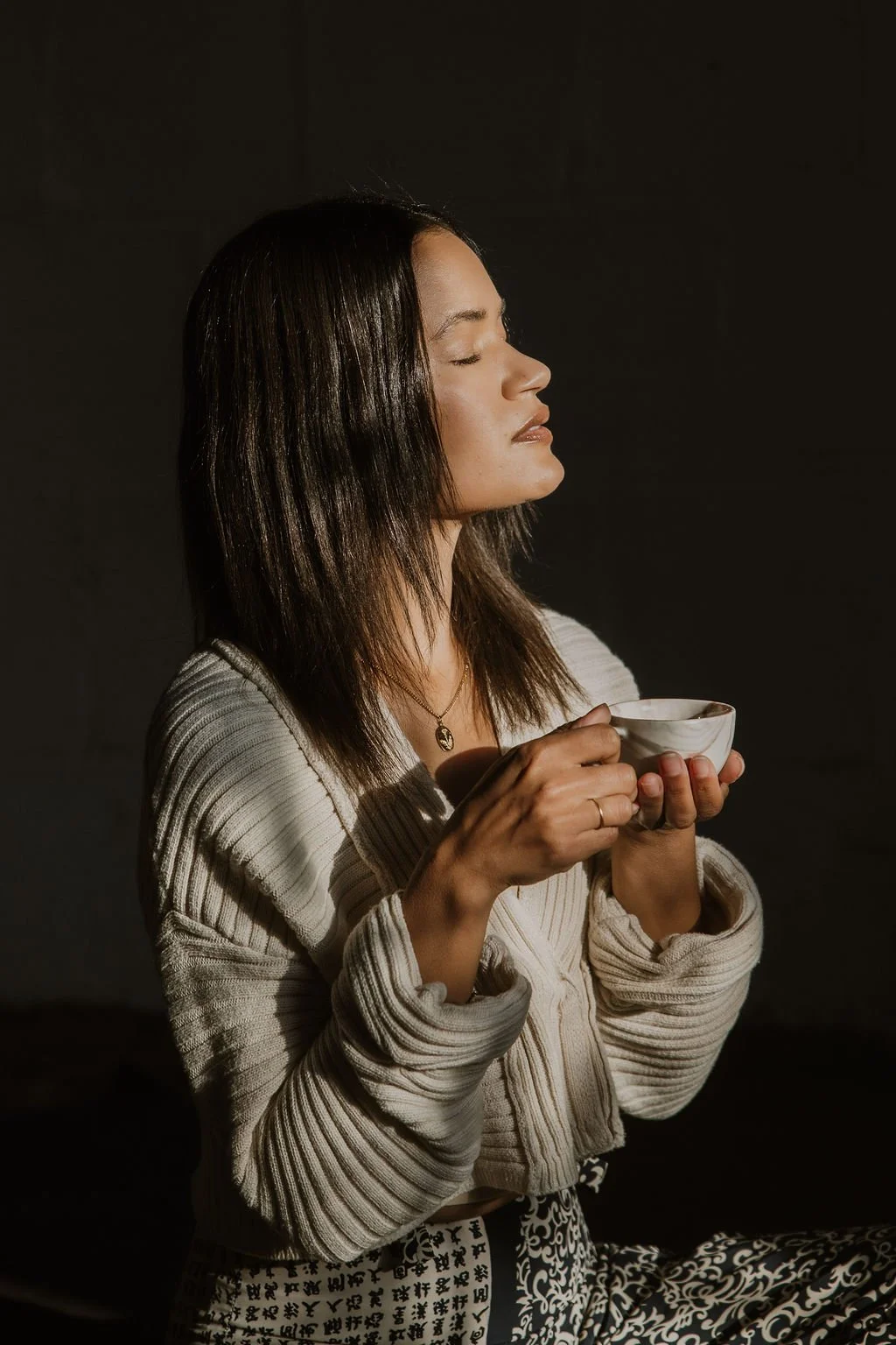 A woman with shoulder-length dark brown hair and closed eyes holds a white ceramic mug with both hands, wearing a beige ribbed cardigan and patterned pants against a dark background.
