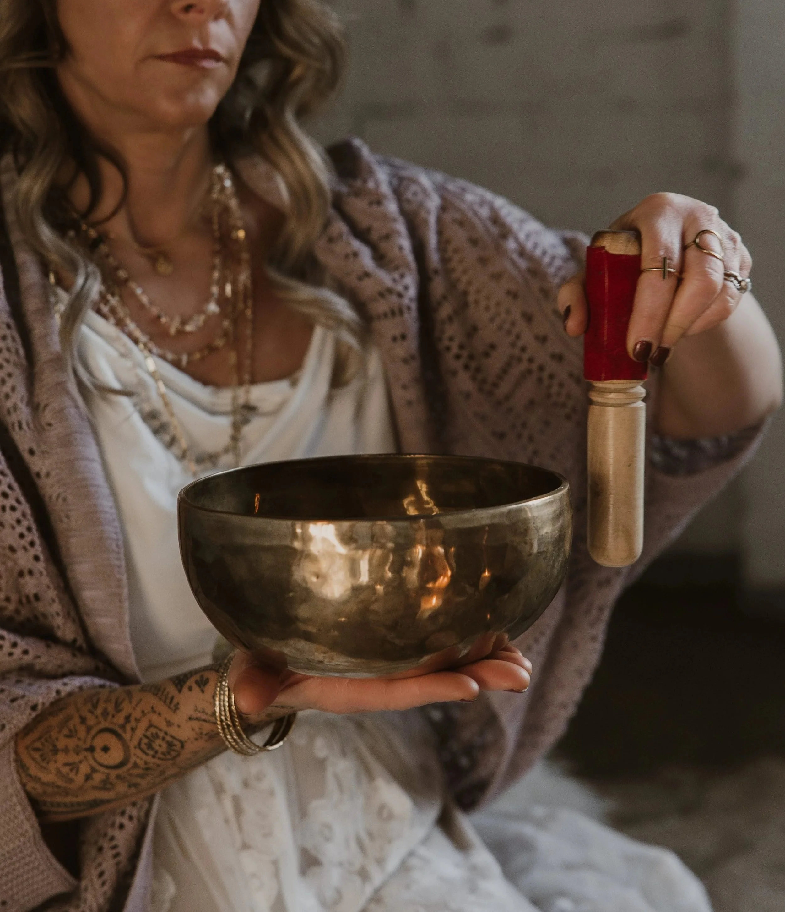 A woman wearing multiple layered necklaces and jewelry, holding a large metallic singing bowl, with her right hand on a wooden mallet, ready to play the bowl.
