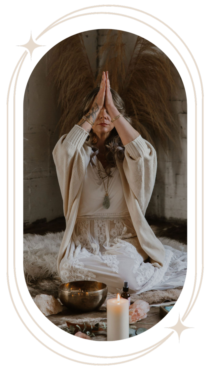 A woman with long hair, wearing a cream-colored cardigan and white lace clothing, sits cross-legged on a fur rug with her hands pressed together in a prayer or meditation pose. She is in a cozy, rustic setting with dried plants behind her and surrounded by candles, crystals, a singing bowl, and essential oil bottles.