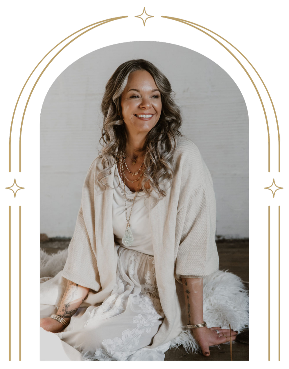 A smiling woman with long wavy hair sitting on a furry white rug, wearing layered necklaces, a light-colored dress, and a light cardigan in a minimalistic setting.