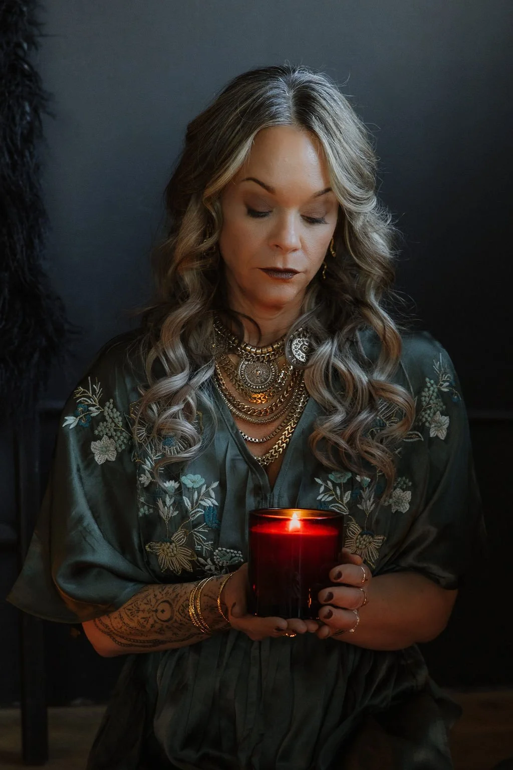 A woman with wavy, blonde hair holding a lit red candle, wearing multiple gold necklaces, a ring, and a dark embroidered blouse, standing against a dark background.