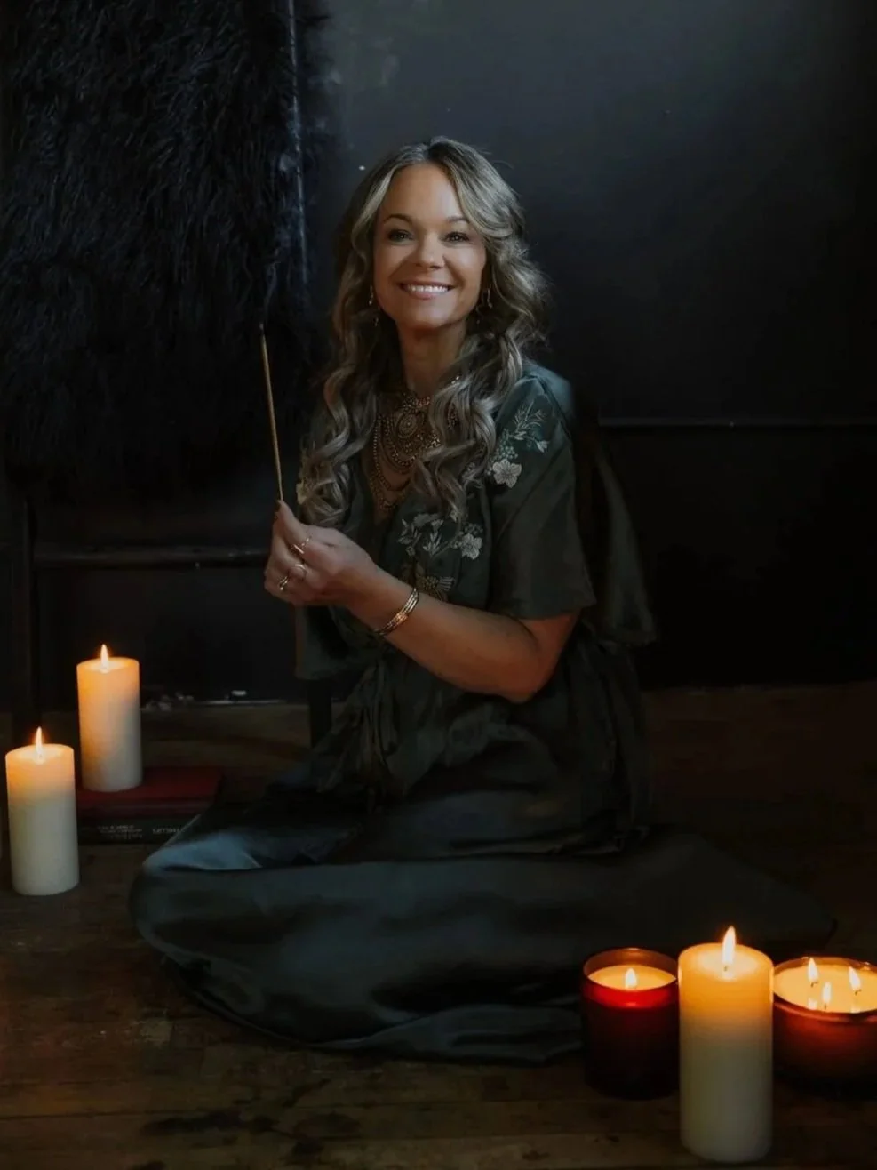 A woman with long curly gray hair, sitting cross-legged on the floor, smiling, surrounded by lit candles, holding a black feather pen, in a dark room with candles and books nearby.