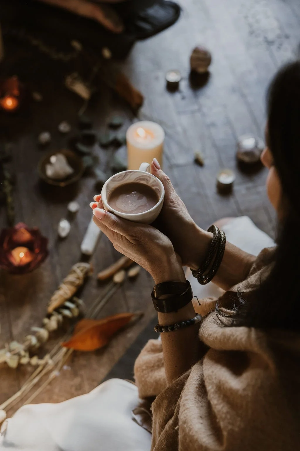 A woman holding a mug of coffee or hot chocolate, surrounded by candles, candles holders, and decorative items on a wooden floor, with a cozy, warm ambiance.