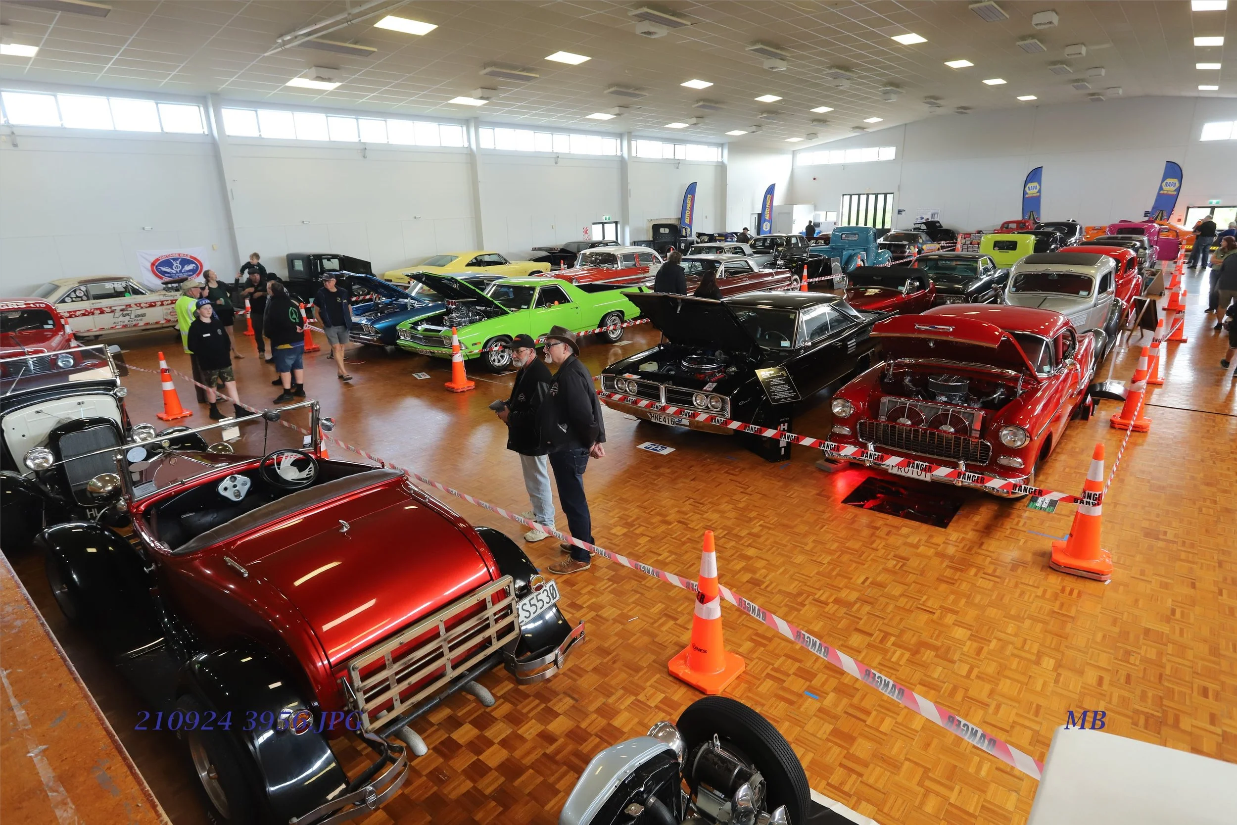 A large indoor car show featuring a variety of vintage and classic cars on display. Visitors are walking around and observing the automobiles, which are roped off with orange and white caution tape. The room has a high ceiling with fluorescent lighting and large windows letting in natural light.