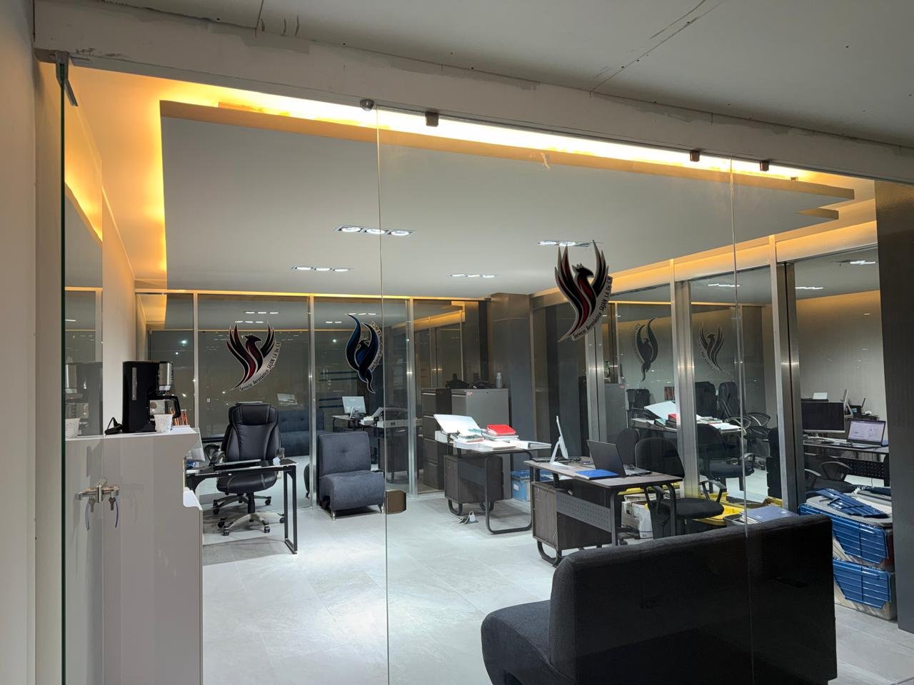 Vista de una oficina moderna con escritorios, sillas de oficina, computadoras y archivadores, vista a través de un muro de vidrio con logotipos en las ventanas.