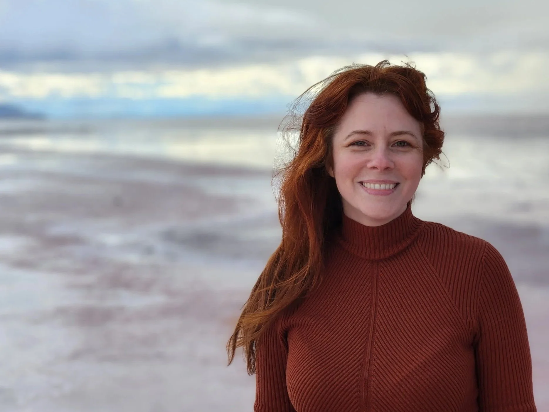 A woman with long red hair smiling on a beach with cloudy skies and ocean waves in the background.