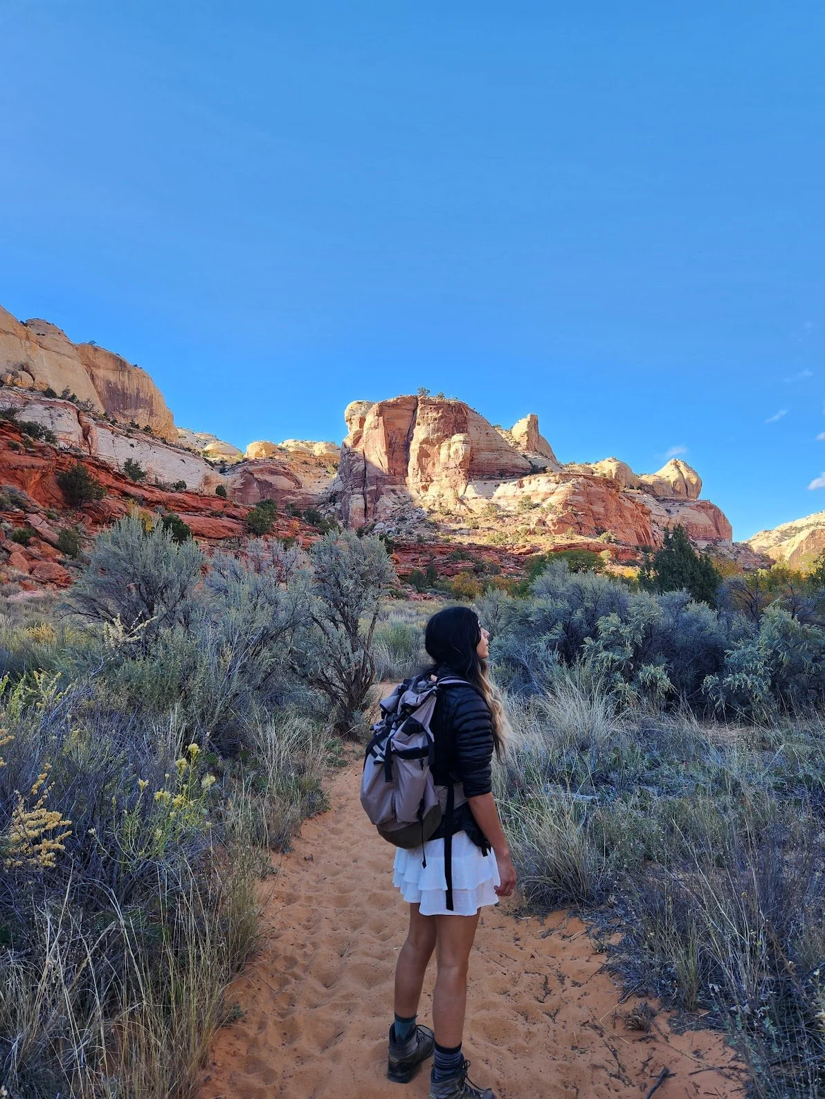 A woman hiking on a dirt trail in a desert landscape with red and white rock formations under a clear blue sky.