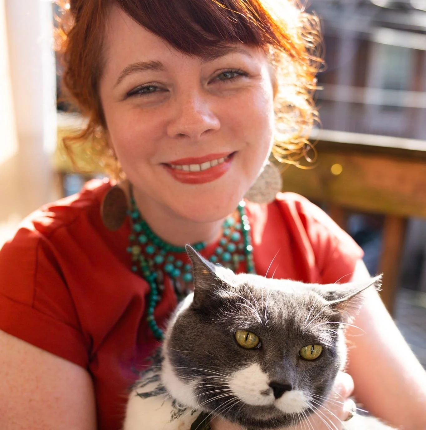 A woman with red hair and a red dress smiling while holding a gray and white cat outside on a balcony in the sunlight.
