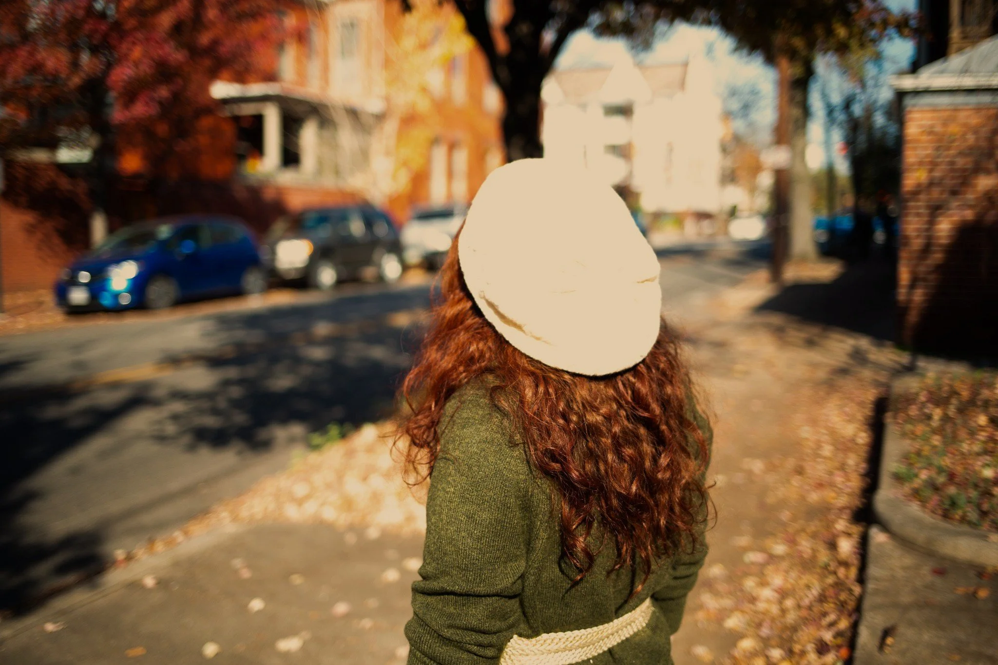 A girl with red curly hair stands on a sidewalk during autumn, wearing a white knit hat and a green sweater. She is facing away from the camera, looking towards houses and parked cars in the distance, with fallen leaves on the ground.