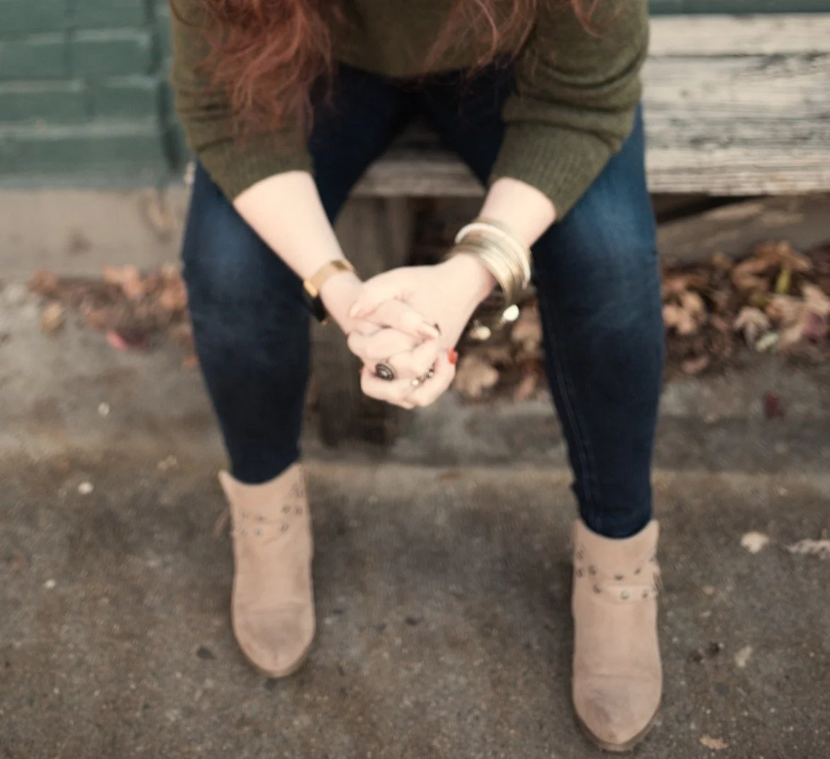 Person sitting on a bench, wearing tan boots, dark jeans, a green sweater, with hands clasped and wearing jewelry, outdoors with fallen leaves.