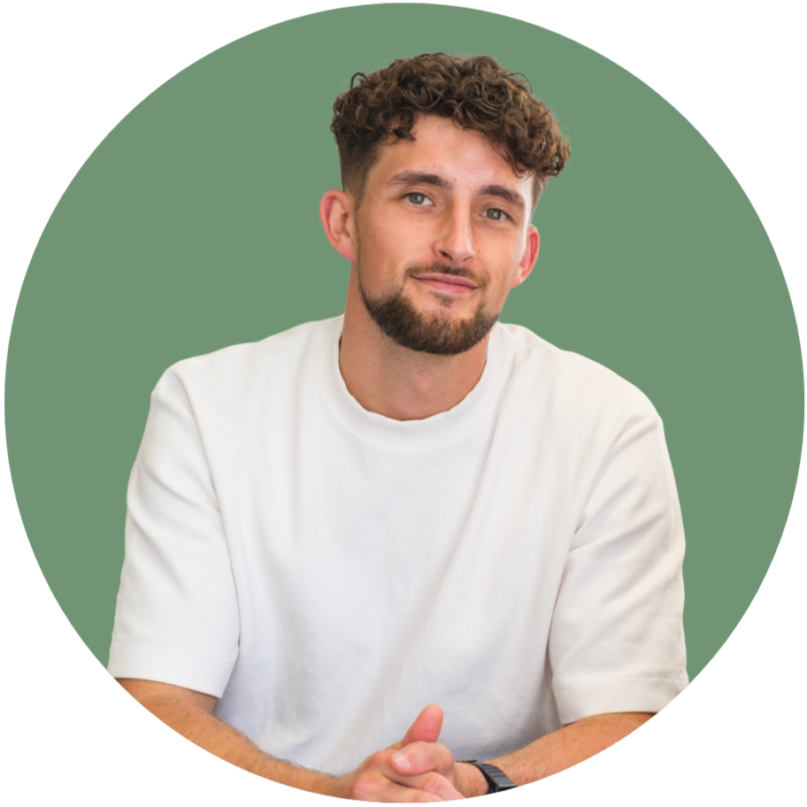 A young man with curly brown hair, a beard, and light skin, wearing a white t-shirt, sitting with his hands clasped in front of him against a green circular background, looking at the camera and smiling slightly.