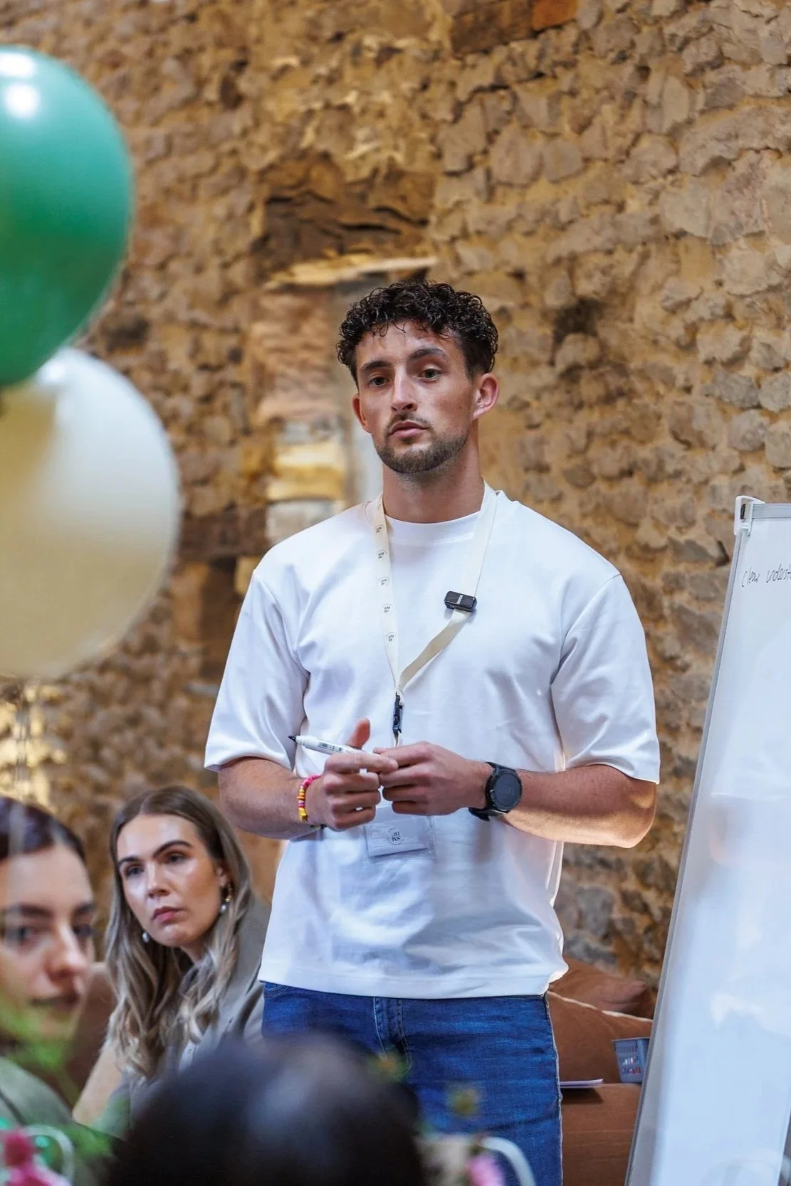 A young man with curly dark hair stands in front of a brick wall, holding a marker, wearing a white T-shirt, a watch, a wristband, and a lanyard with a microphone clipped on. He appears to be giving a presentation or lecture to a group of people, some of whom are visible in the foreground, with a whiteboard nearby.