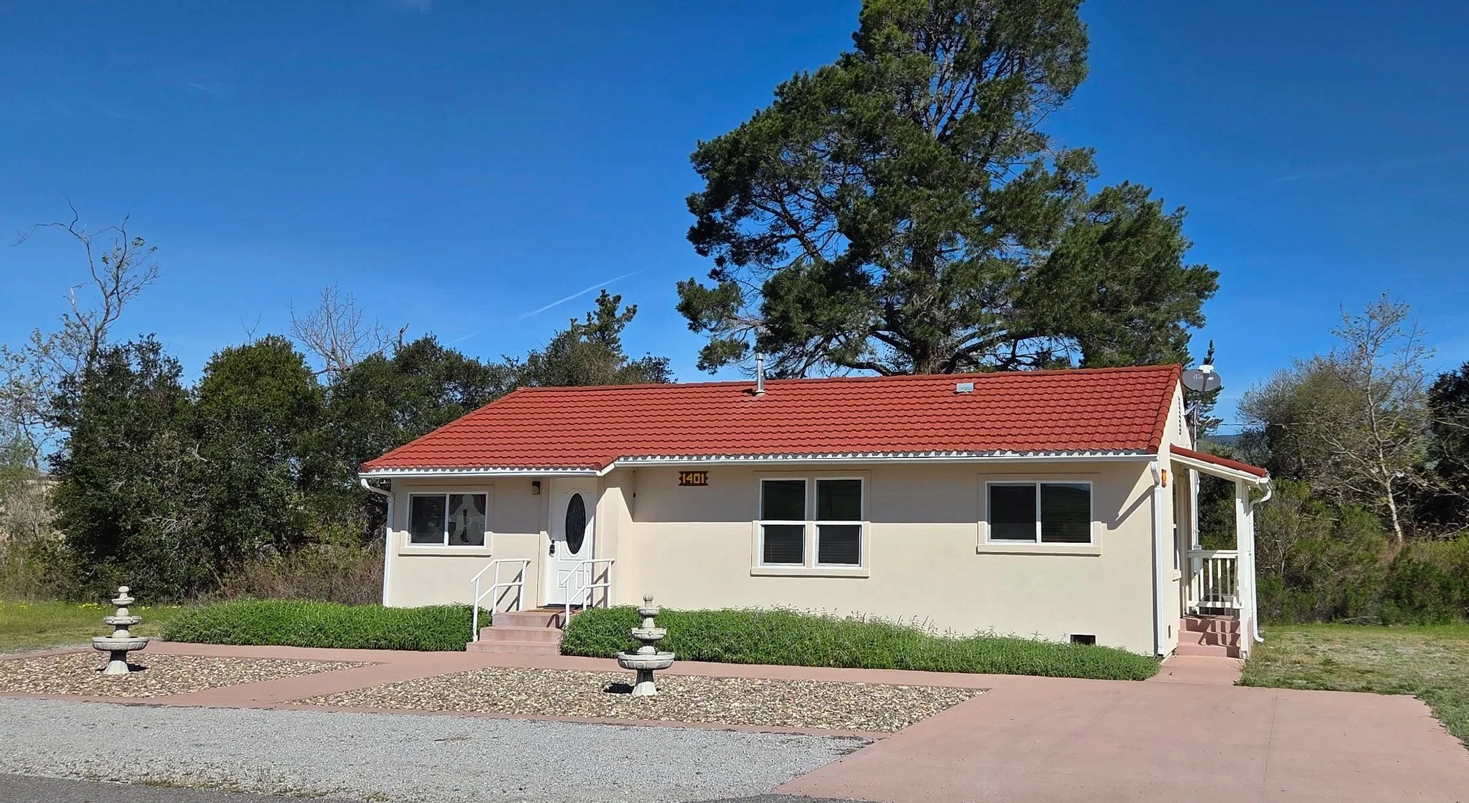 Single-story house with a red tile roof, white exterior walls, front stairs, surrounded by a well-maintained lawn and bushes, with a large tree in the background and a bright blue sky.