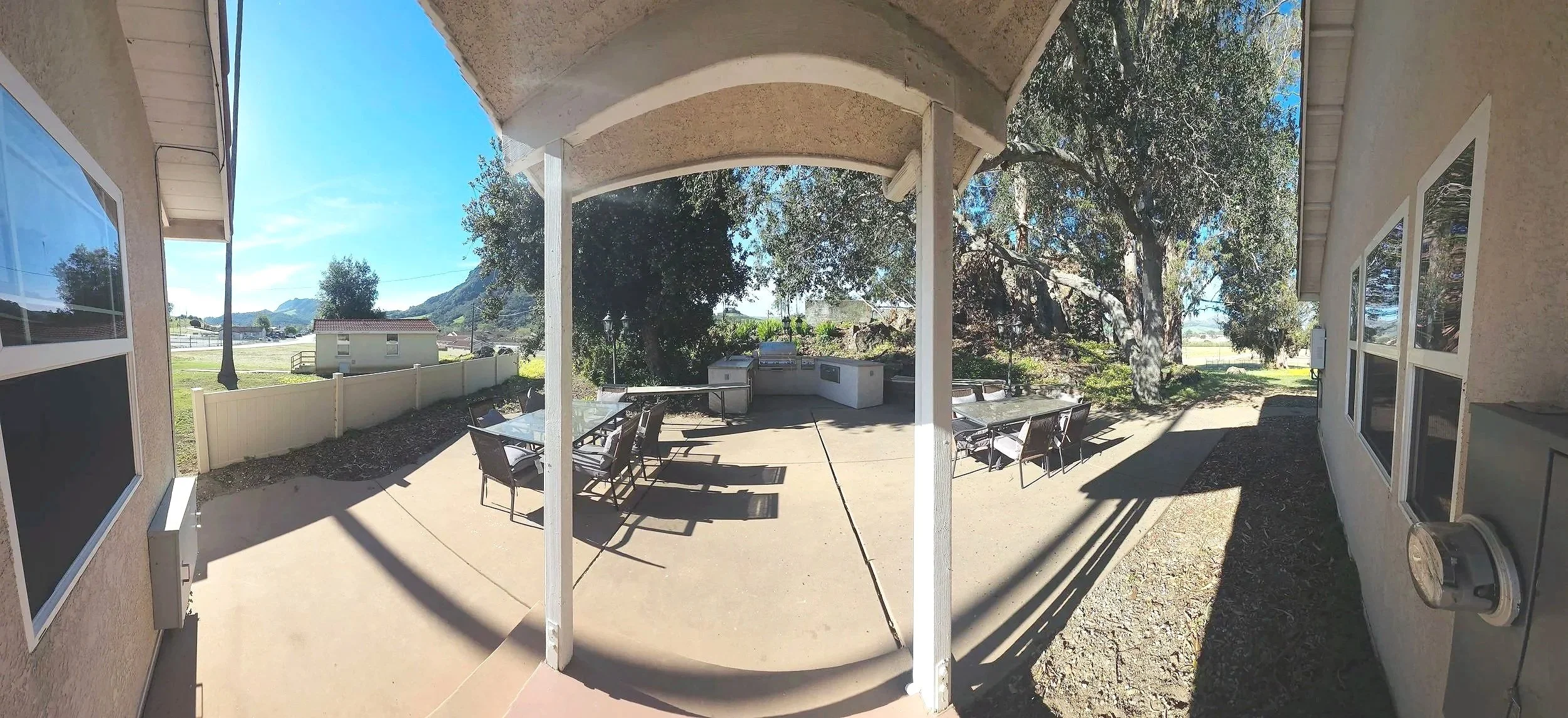 A spacious outdoor patio with tables and chairs, surrounded by trees and mountains in the distance. The patio has a covered section and sunny areas, with a barbecue grill and a low fence separating it from the grassy landscape.