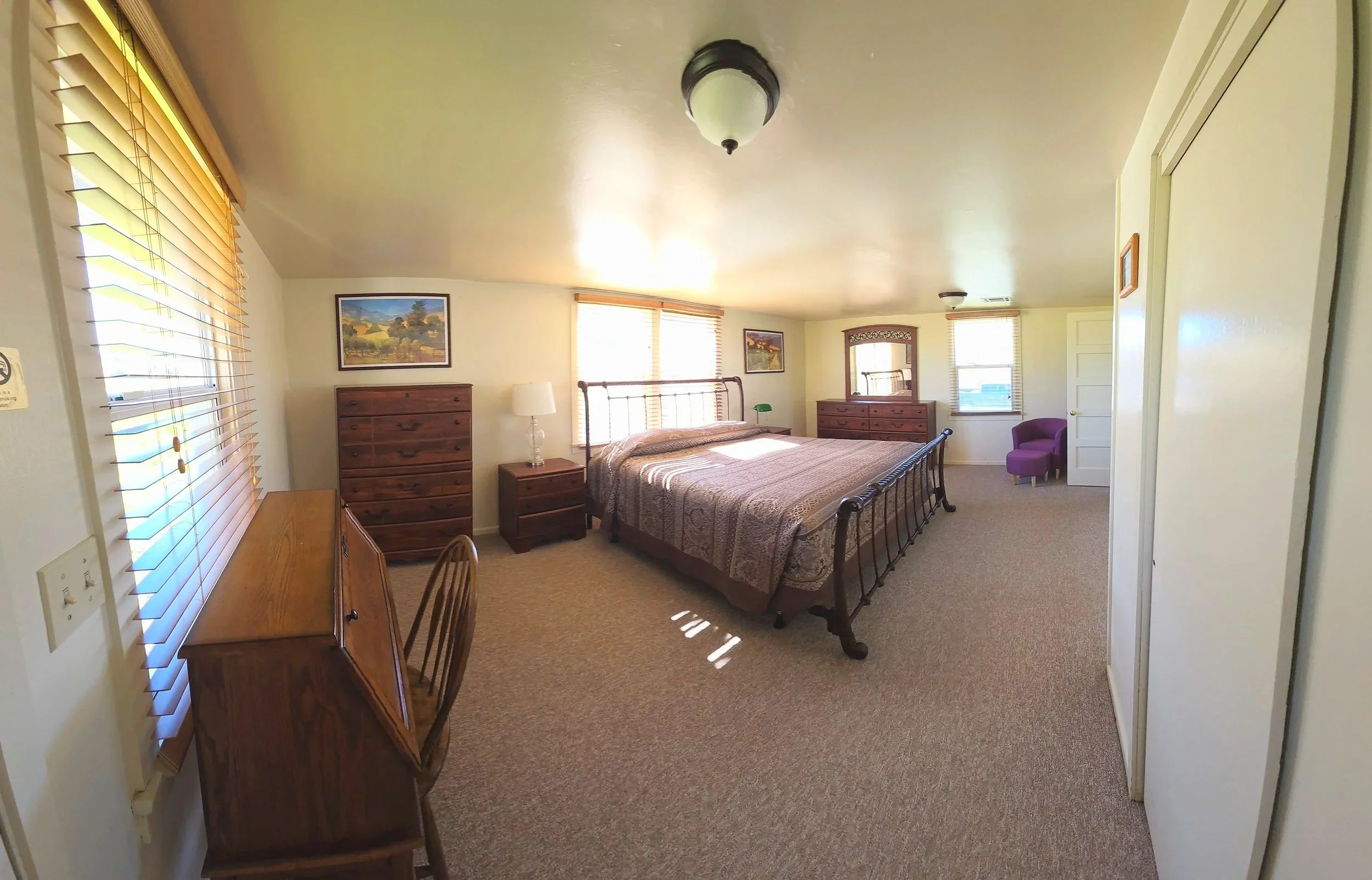 Spacious bedroom with a large bed, wooden furniture, and a purple armchair, illuminated by natural light through multiple windows.