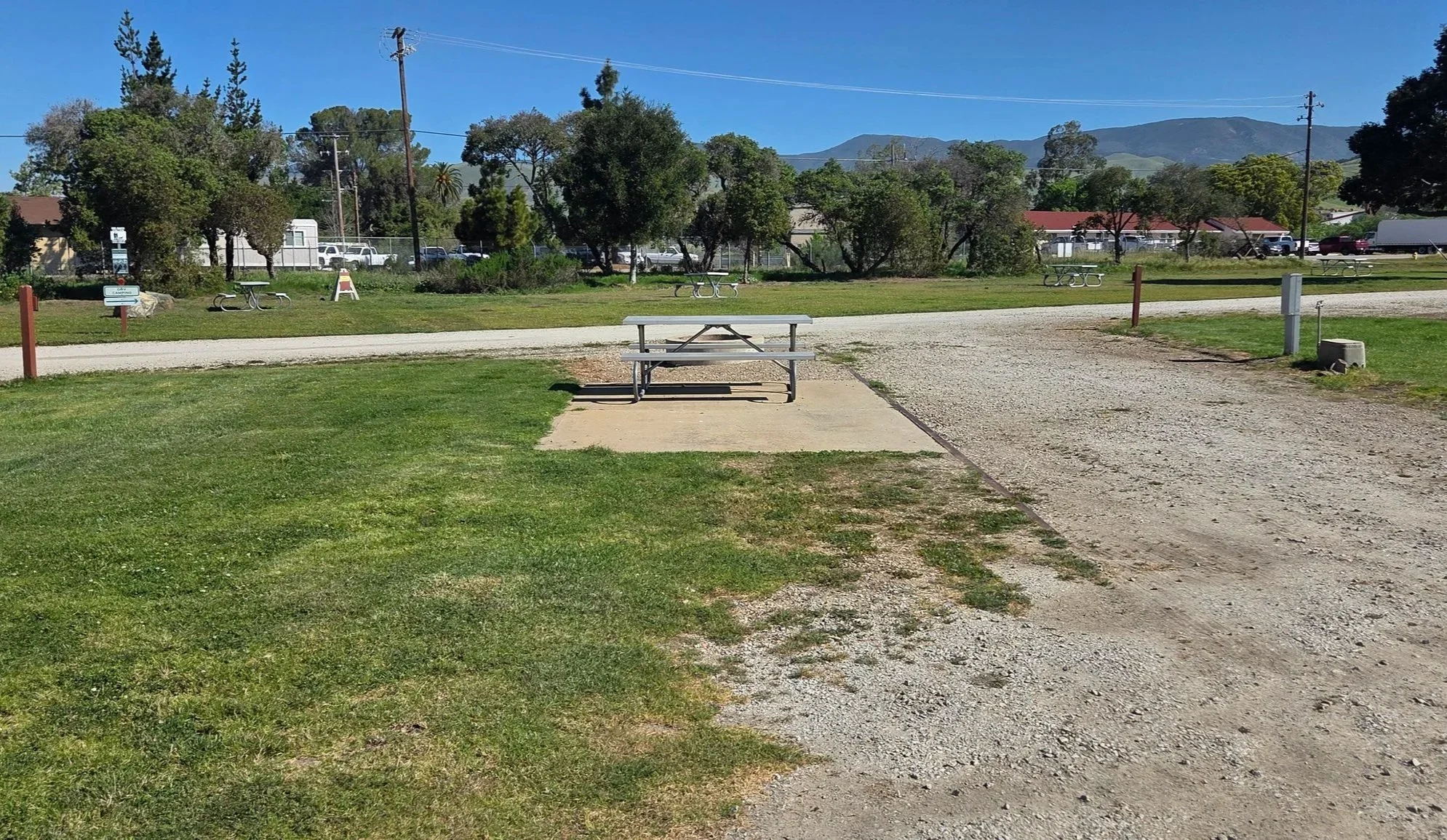A park with a picnic table on a concrete pad, surrounded by grassy areas and dirt pathways, with trees and mountains in the background under a clear blue sky.