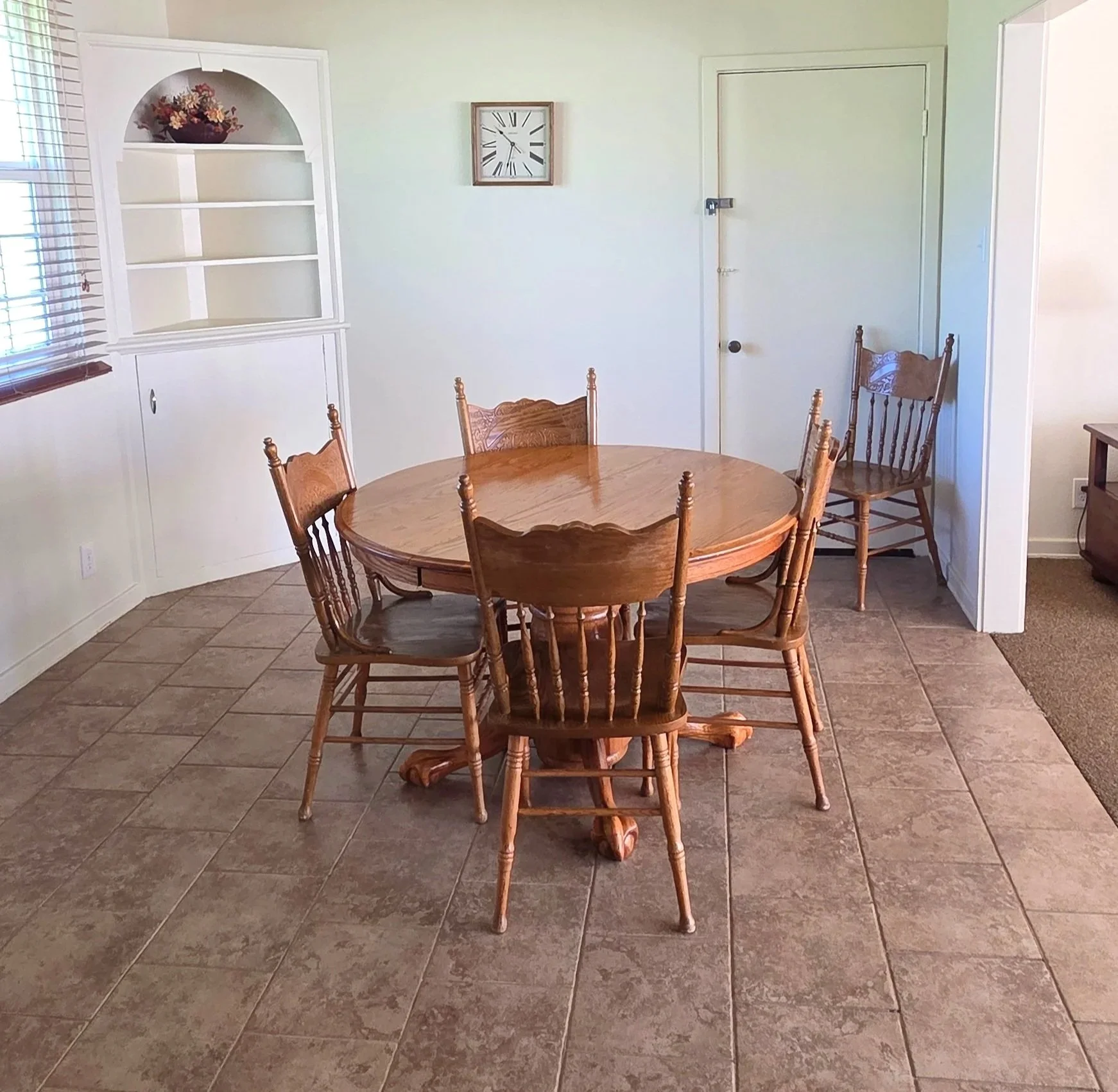 A round wooden dining table with six matching chairs in a room with beige tiled floor and light green walls. A white built-in bookshelf with decorative flowers is on the left. A white wall clock is on the back wall, and there is a closed white door in the back right corner.