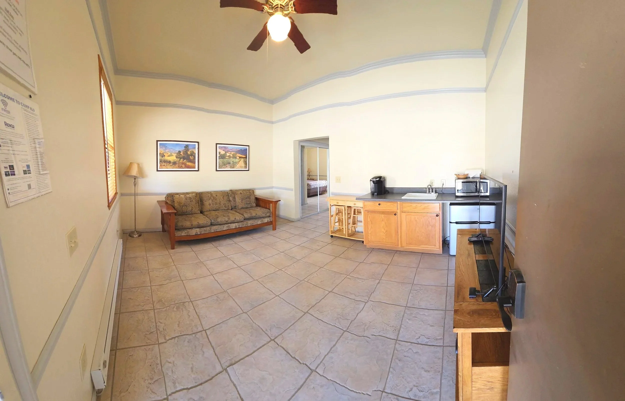 A small living area with a patterned sofa, a floor lamp, two landscape paintings on the wall, a ceiling fan, a kitchenette with a sink, microwave, coffee maker, small refrigerator, and wooden cabinets, tile flooring, and a door leading to a bedroom.