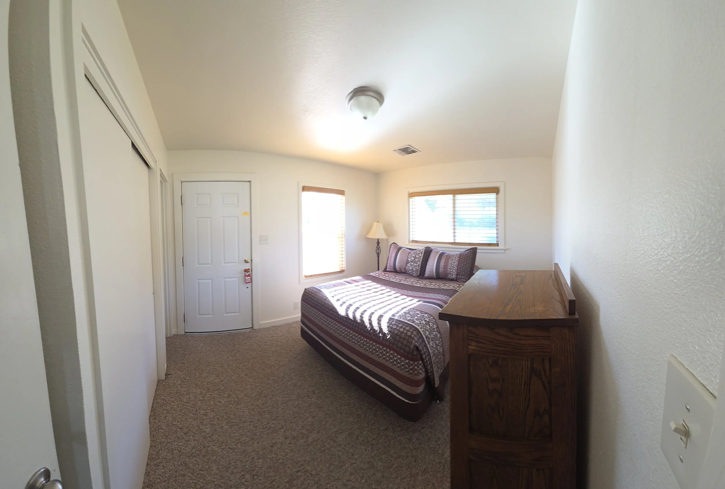 Interior of a bedroom with white walls, a bed with a patterned quilt and pillows, two windows with wooden blinds, a wooden dresser, a floor lamp, and a ceiling light fixture.