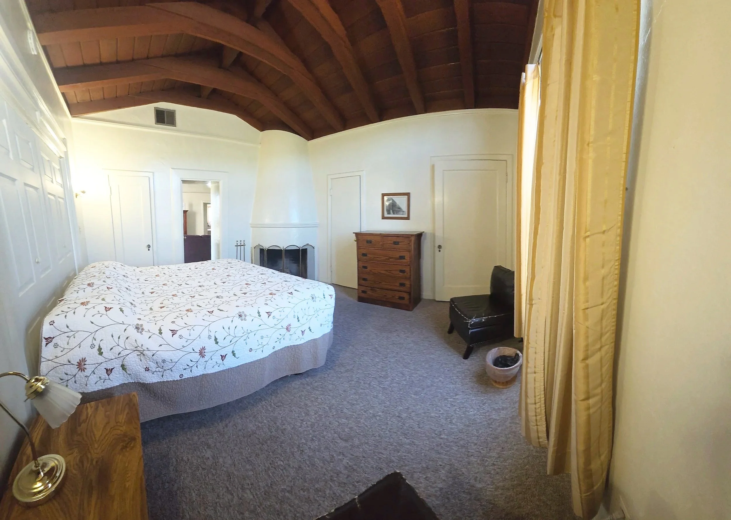 A bedroom with a vaulted wooden ceiling, a window with yellow curtains, a large bed with a floral quilt, and a wooden nightstand with a small lamp.