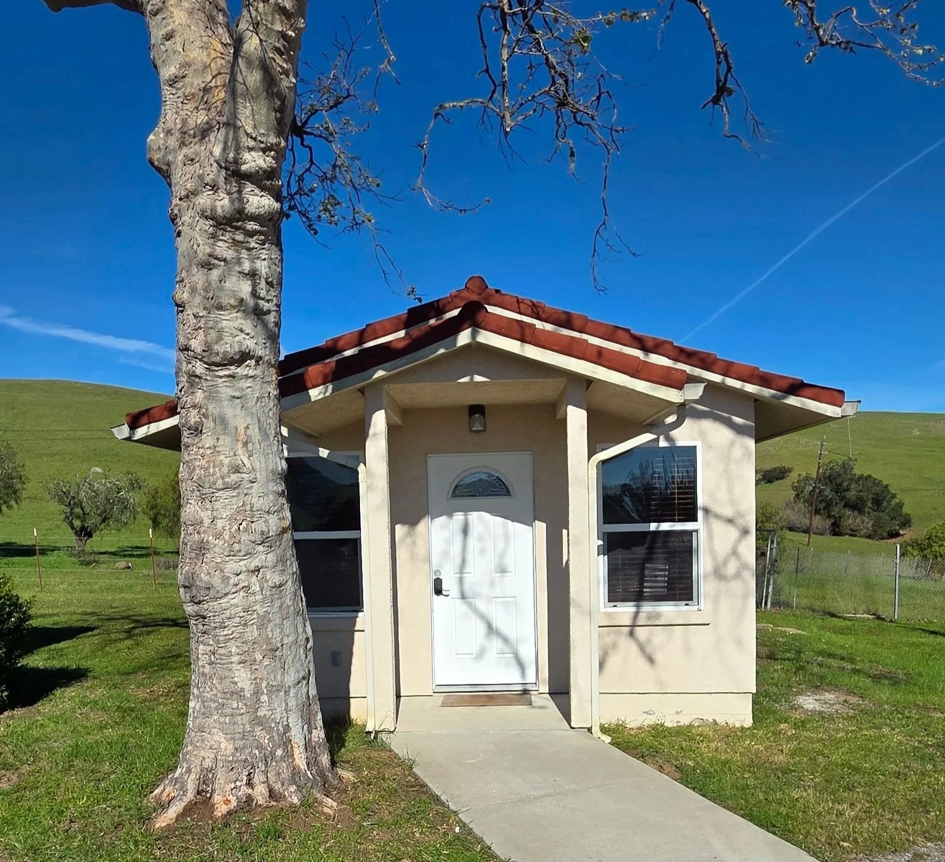 Small beige house with a red tile roof, white door, and two large windows, surrounded by green grass and a large tree in front. Hills and blue sky in the background.