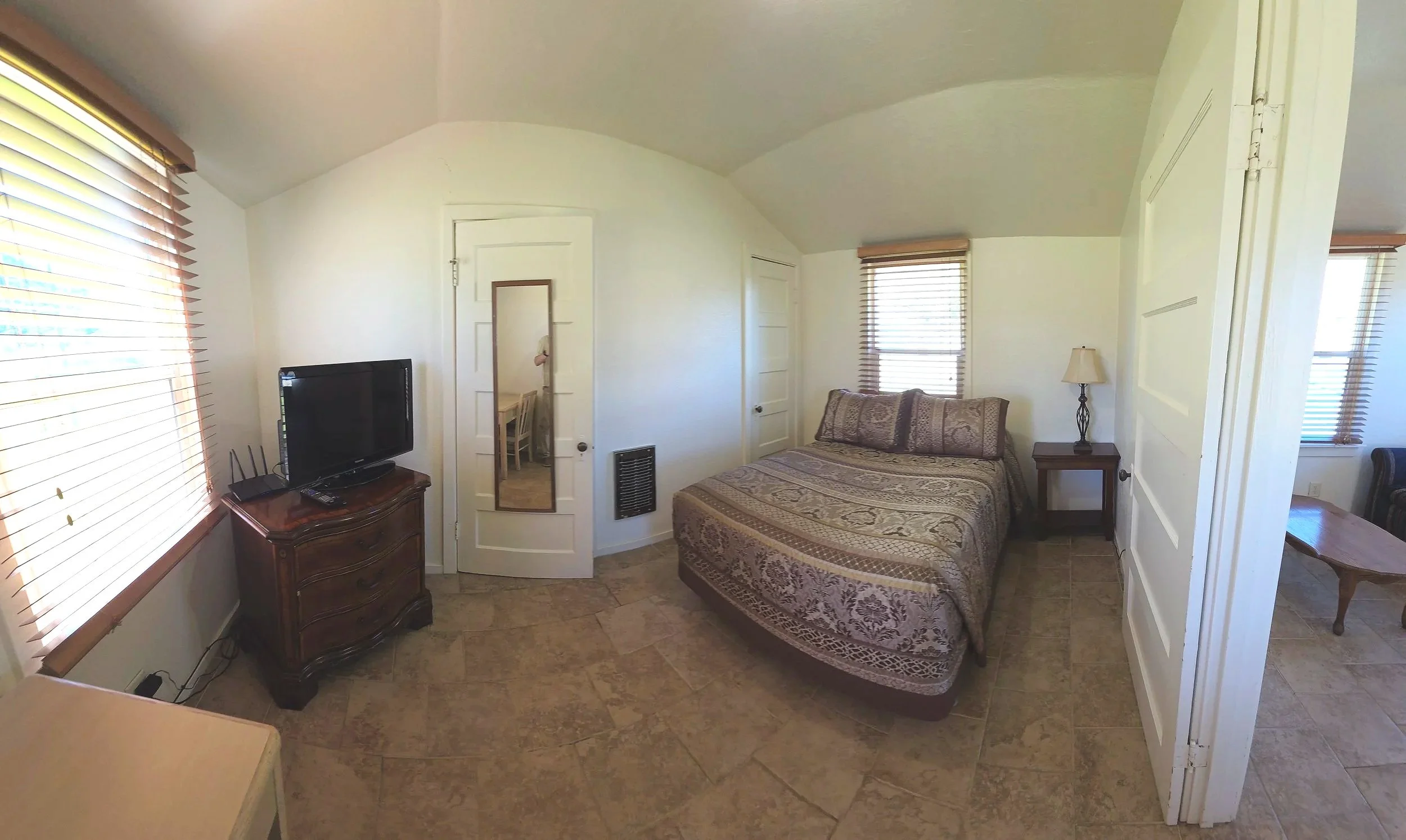 Small bedroom with a bed, side table with lamp, window with blinds, TV on a wooden dresser, white walls, tiled floor, mirror on door.