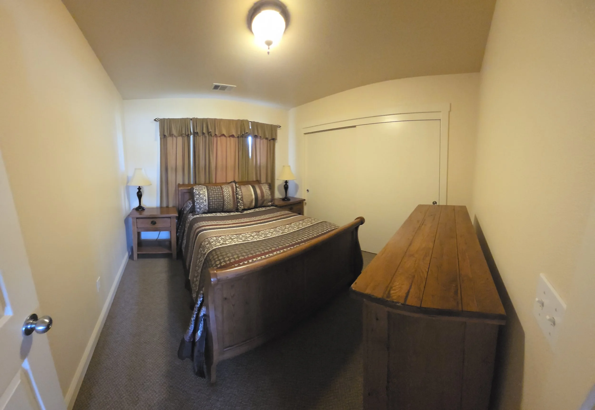 A small bedroom with a wooden bed covered in pattern bedding, two nightstands with lamps, a window with curtains, and a wooden dresser.