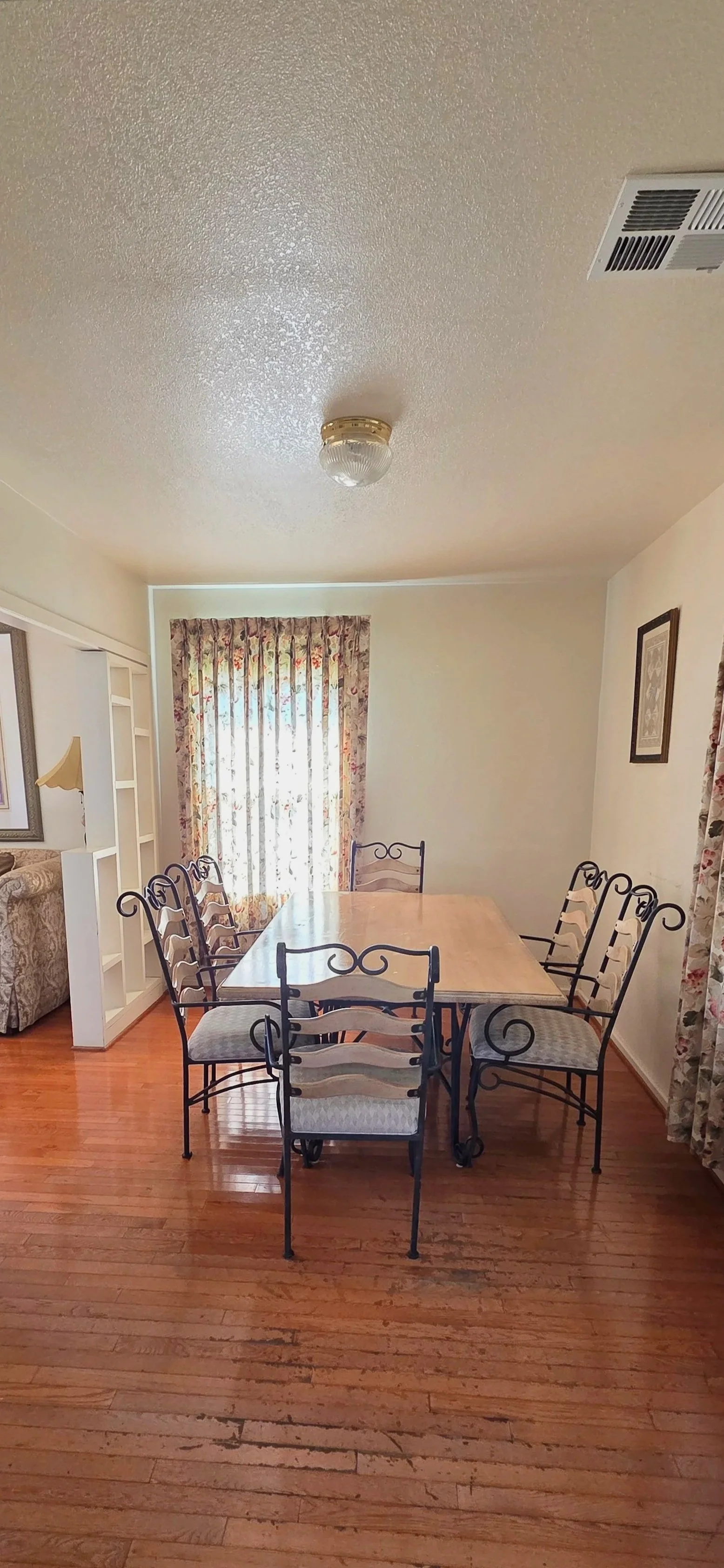 A dining room with a wooden table and six metal chairs with cushioned seats. There are floral curtains on a window and a closed curtain on another window. A partial view of a living room with a floral armchair and framed artwork on the wall can be seen.