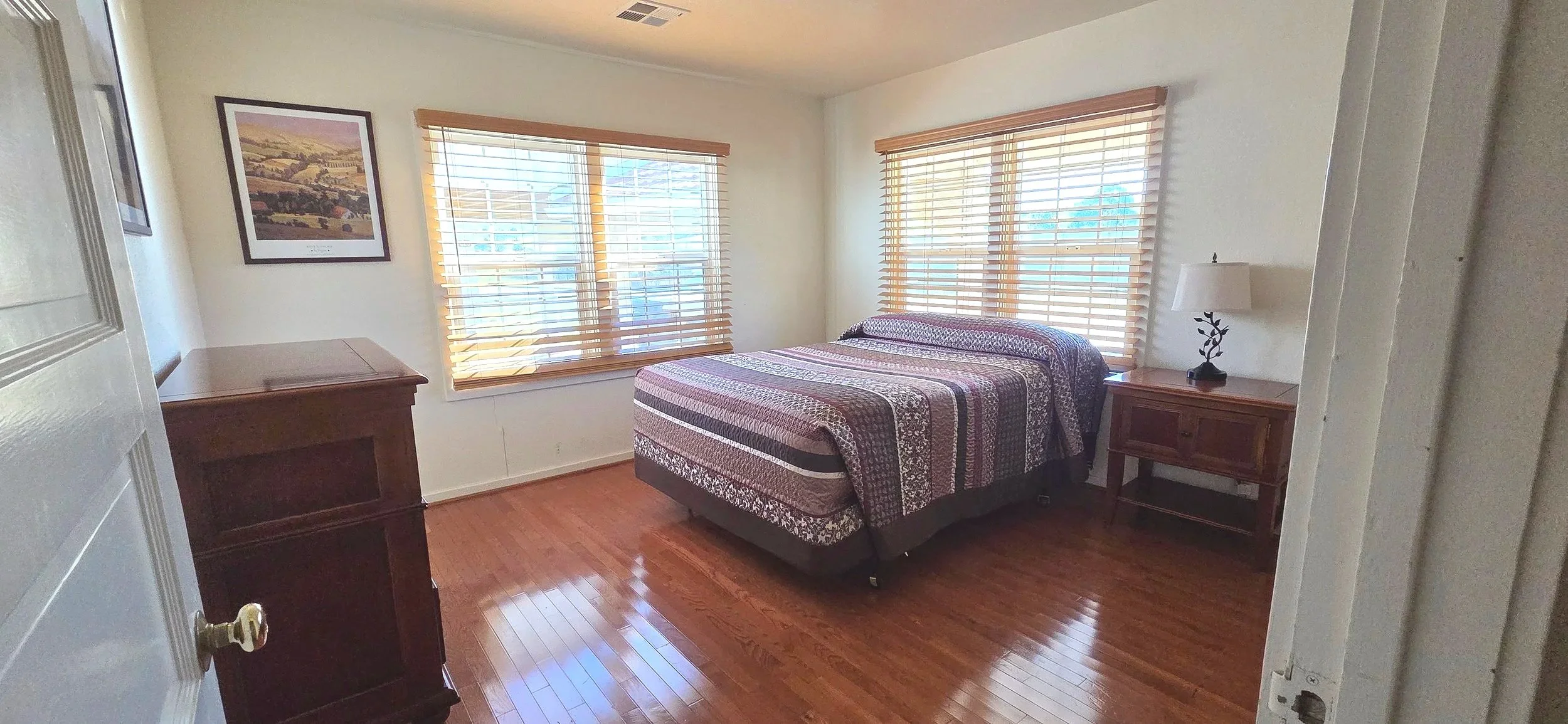 A bedroom with wooden flooring, a bed with a patterned quilt, wooden bedside tables, a lamp, and large windows with blinds letting in natural light.