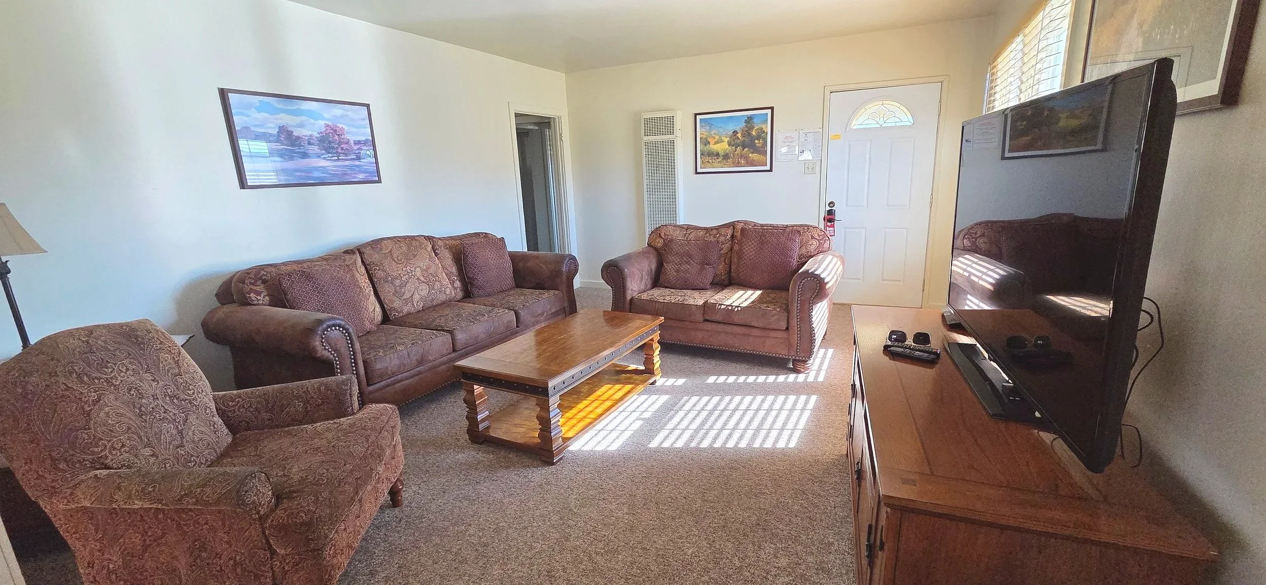 Living room with brown upholstered sofa and armchair, wooden coffee table, flat-screen TV on wooden stand, wall art, and sunlight through window blinds.