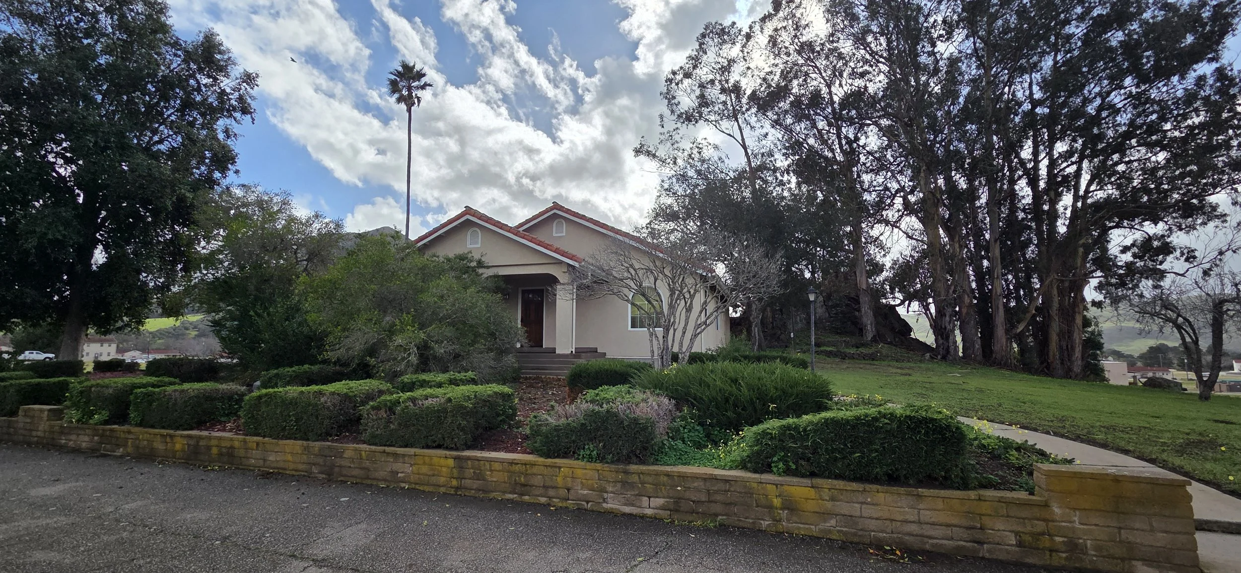A beige house with a red tiled roof, surrounded by trees, bushes, and a manicured lawn, with a sidewalk in front and a cloudy sky overhead.