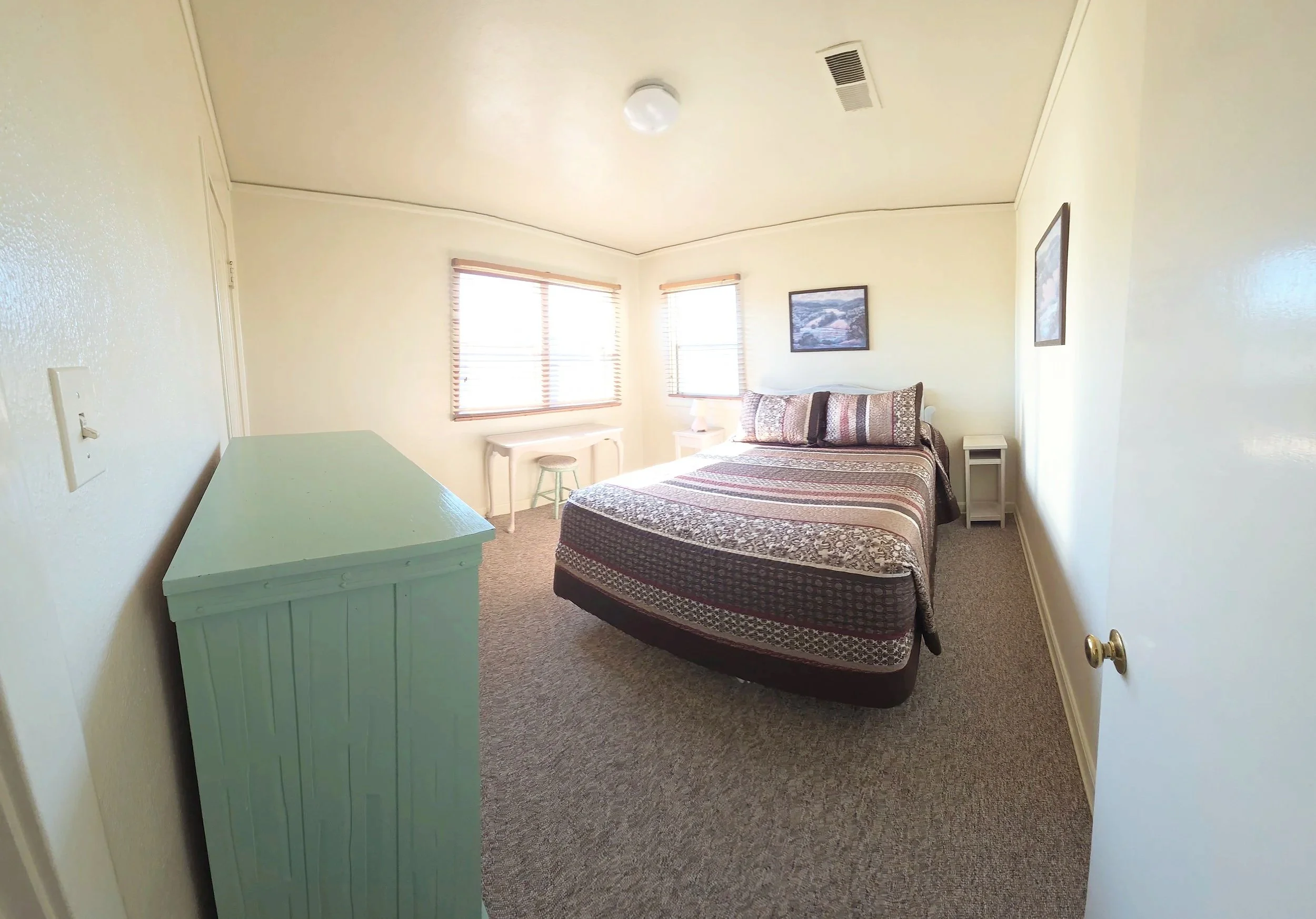 A bedroom with a bed, nightstands, a small desk, and windows with blinds, illuminated by natural light.